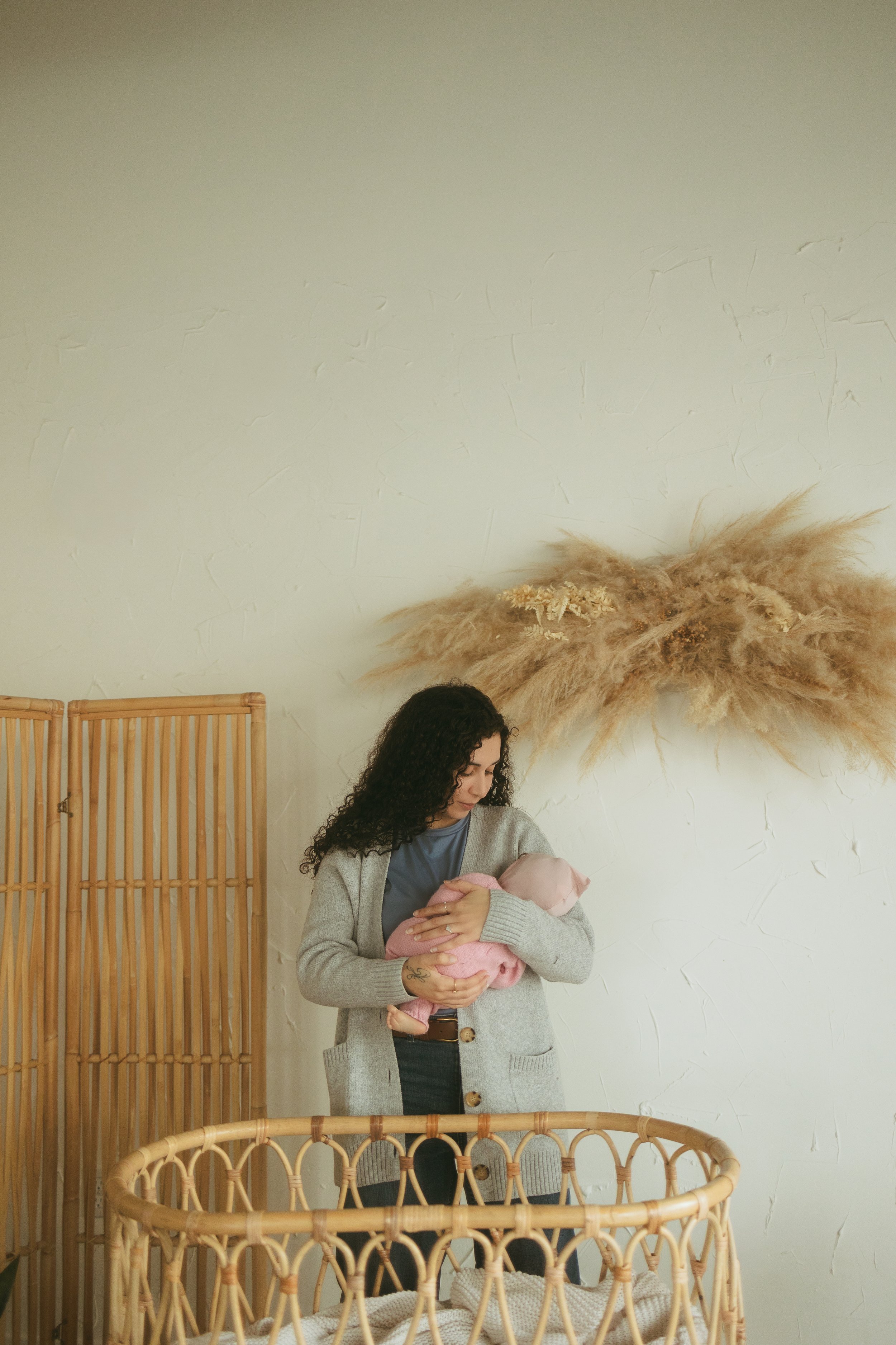 Woman holding a baby in a nursery with rattan furniture and beige pampas grass on the wall.