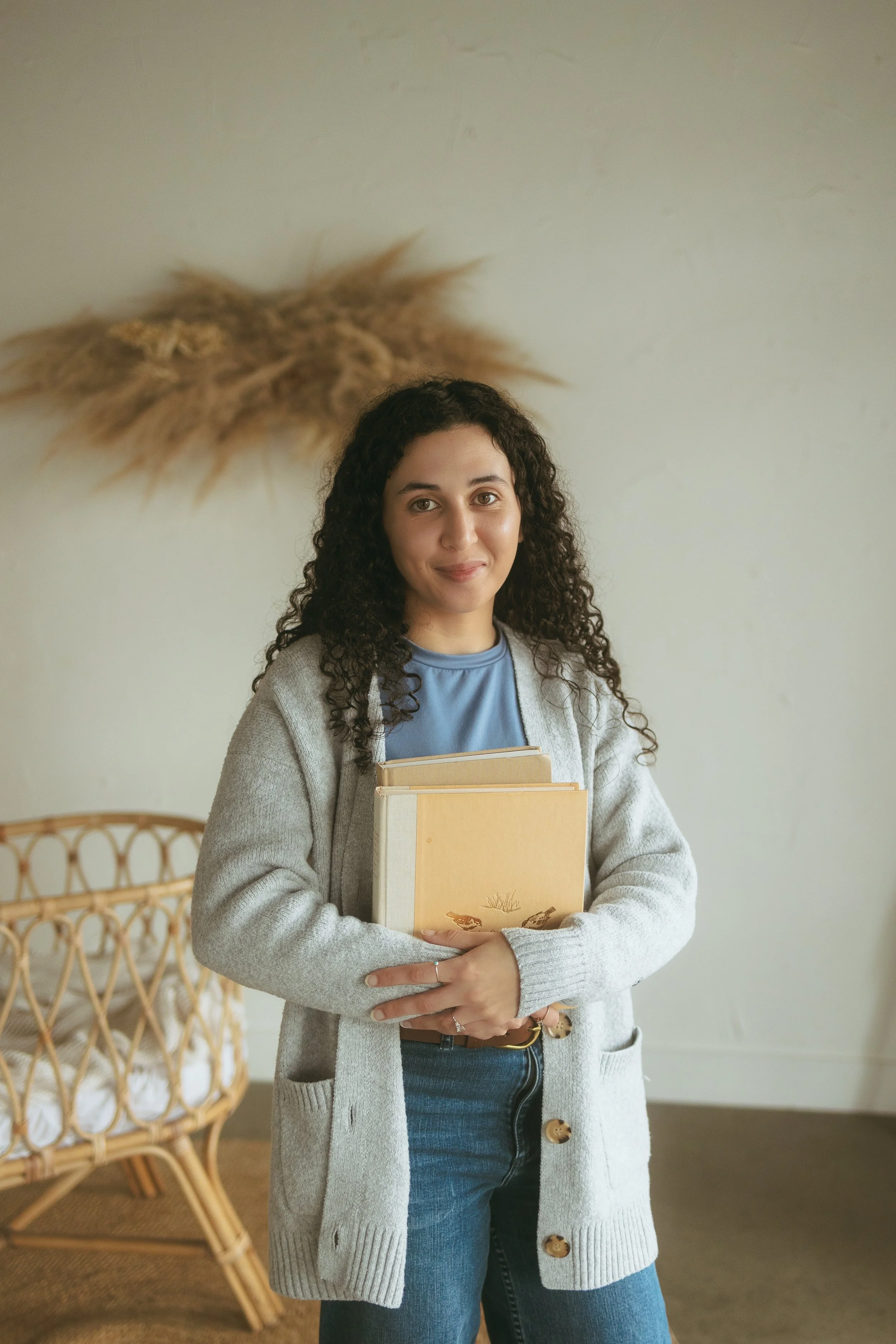A woman with curly dark hair holding books, standing in a room with a wicker chair and a wall decoration in the background.