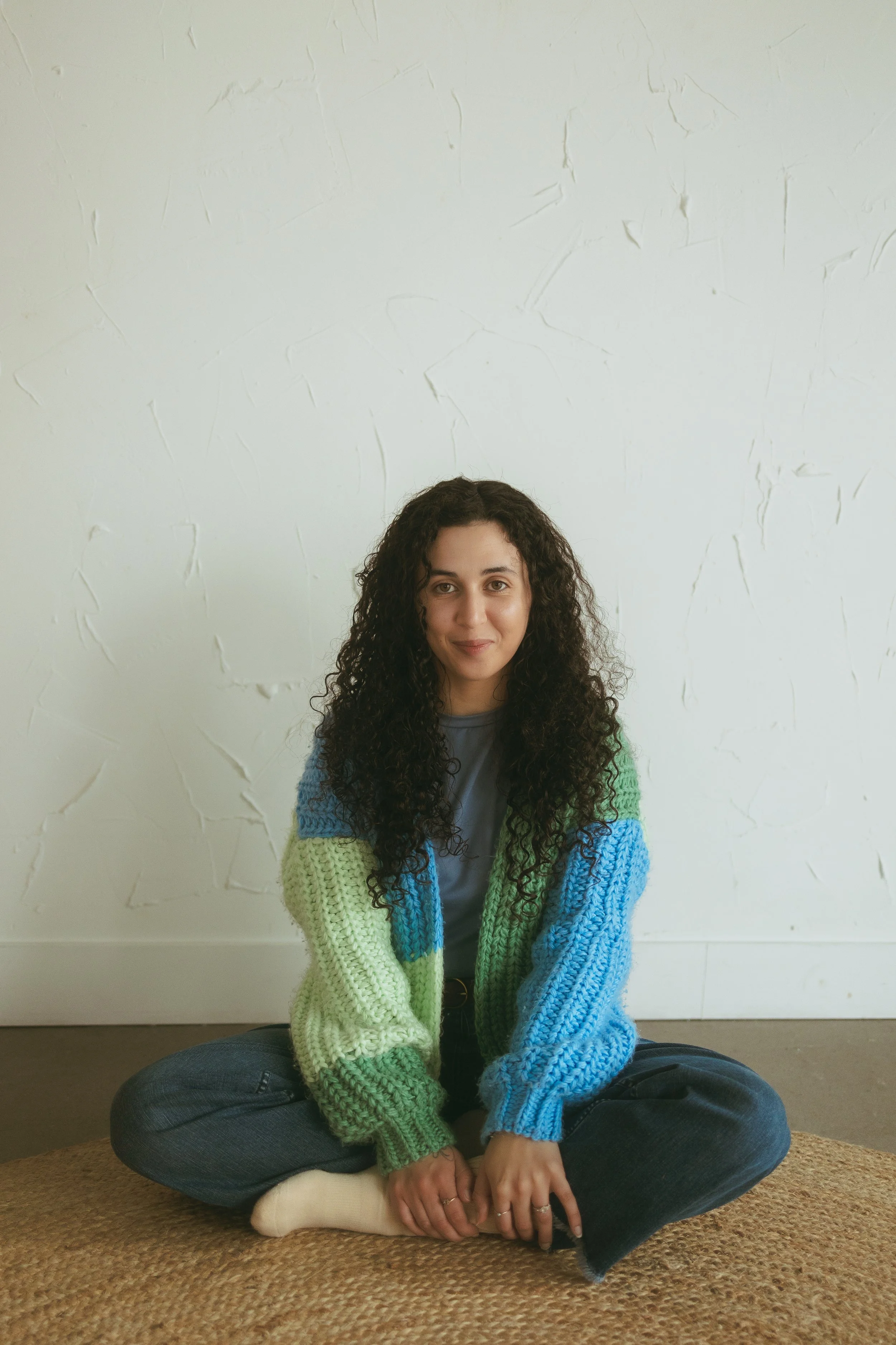 A woman with long curly hair sitting cross-legged on a woven rug against a textured white wall, wearing a multicolored knitted cardigan and a grey t-shirt.