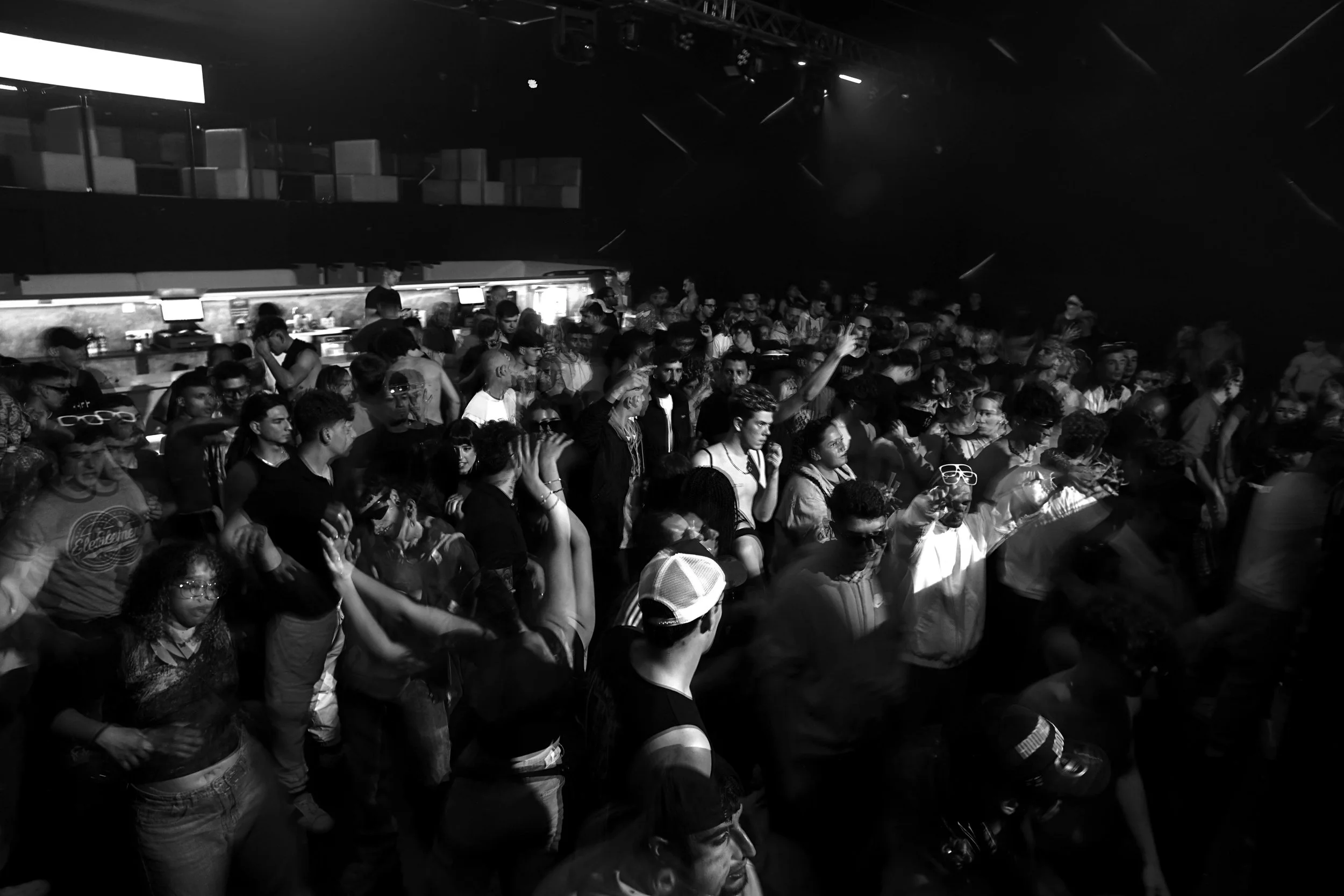 Black and white photo of a crowded indoor dance club with many people dancing and socializing under dim lighting and a bar counter in the background.