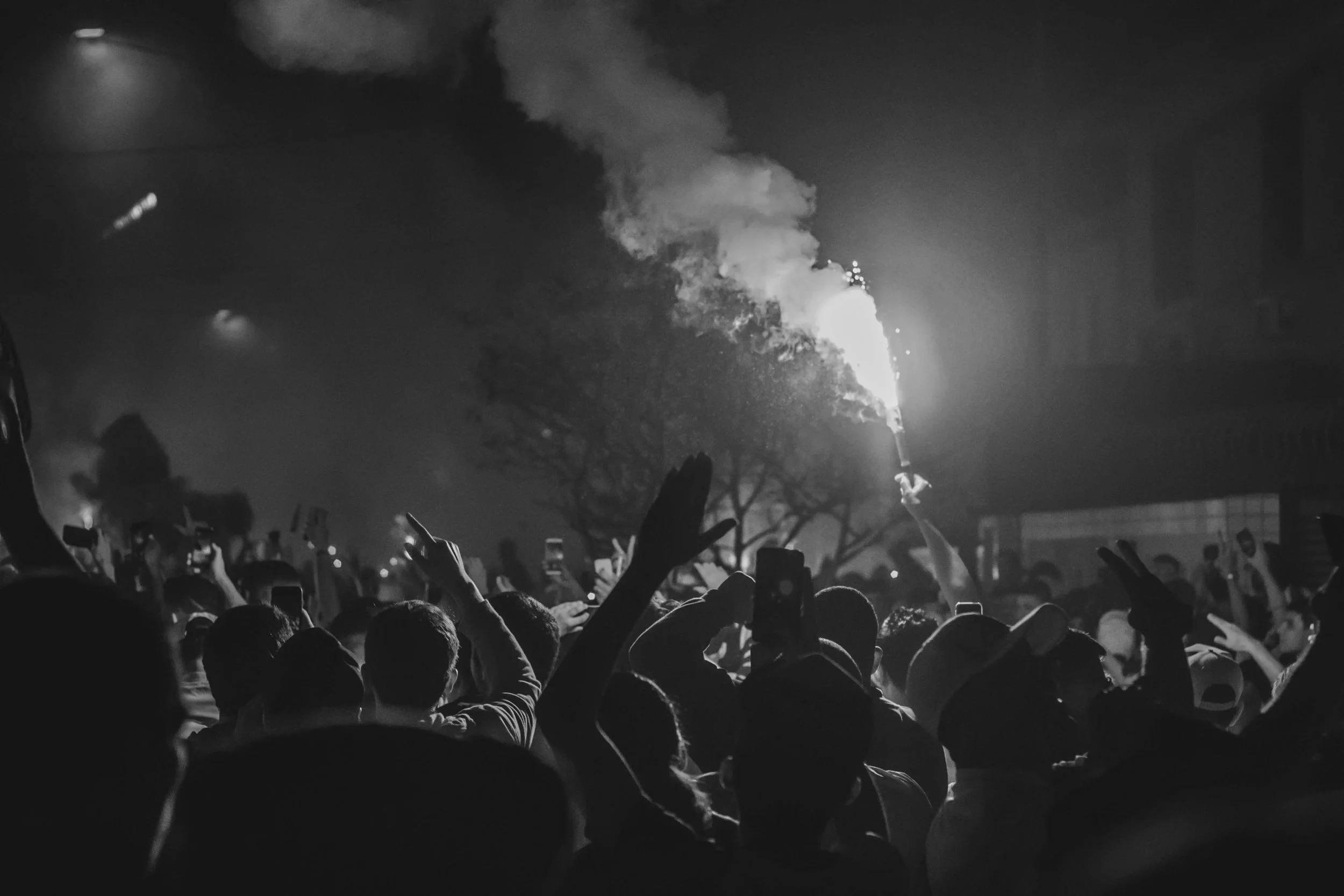 Crowd of people at night holding up phones and a flare, with smoke rising in the air.