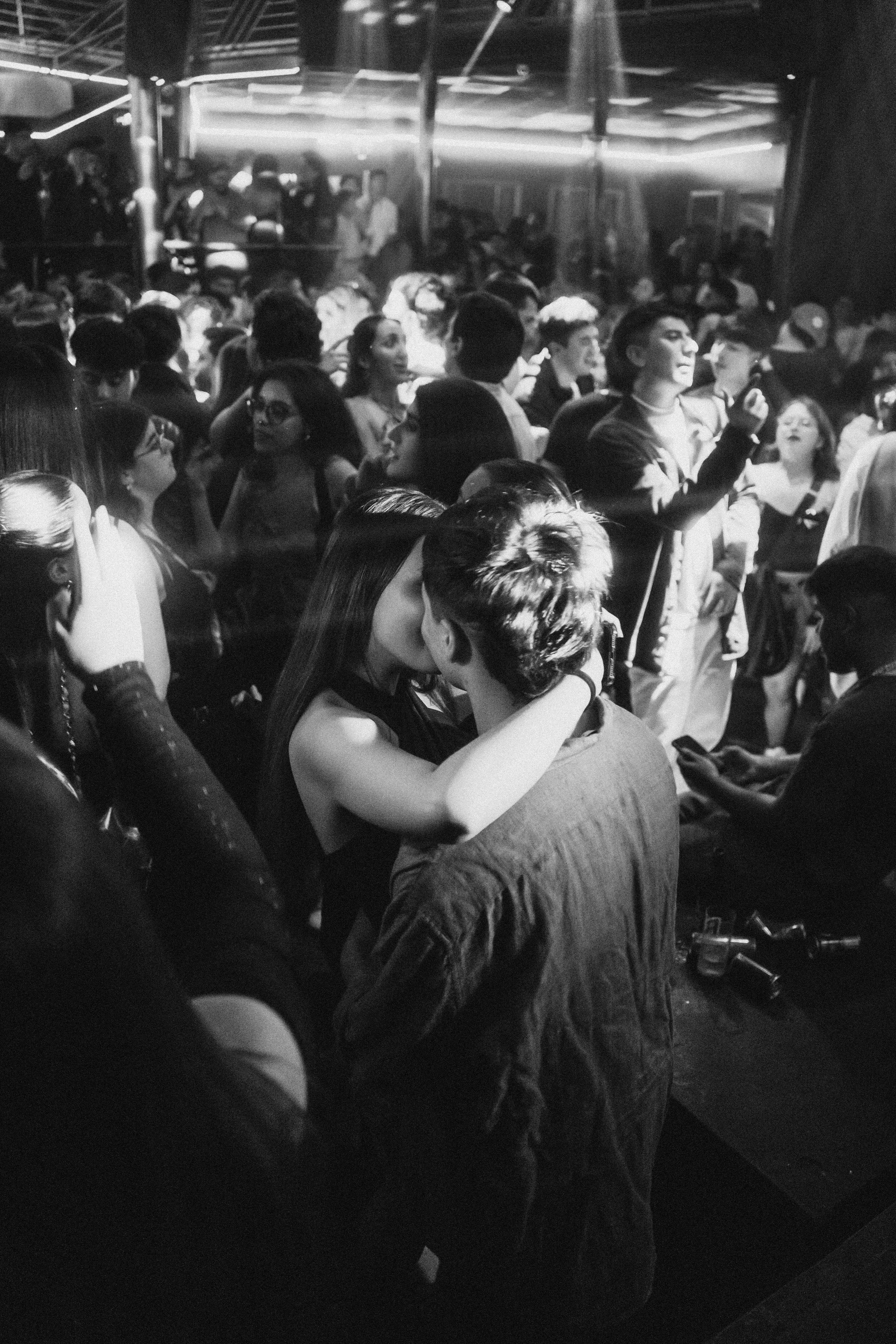 A crowded nightclub dance floor with couples kissing and people socializing, illuminated by dim lighting and surrounded by a lively atmosphere.