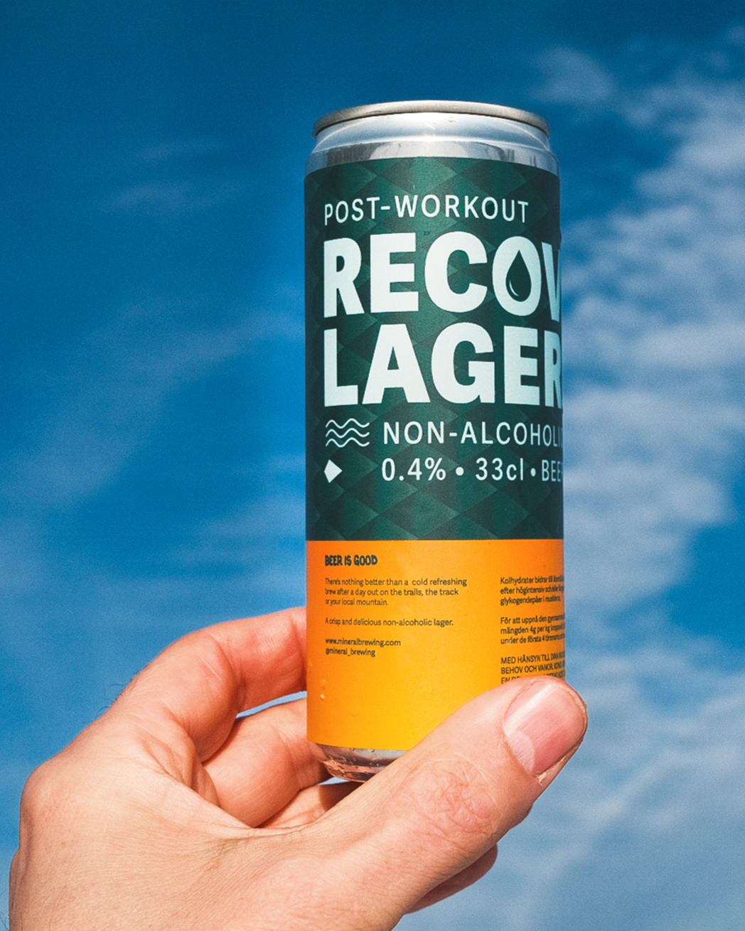 A hand holding a can of 'Post-Workout Recovery Lager'  against a blue sky with clouds.