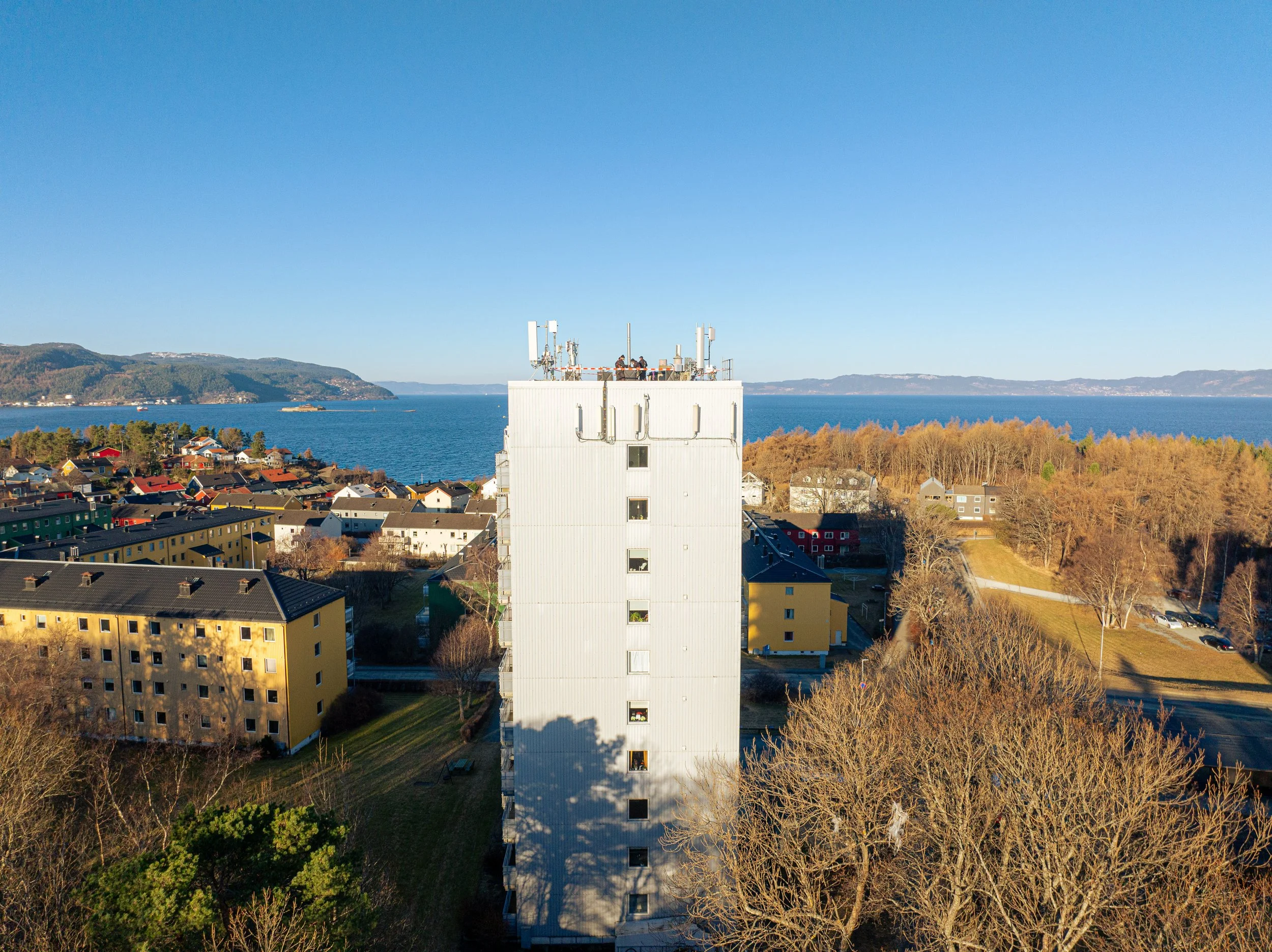 Utsikt over en by med bolighus, en høy bygning med antenner på taket, og sjø med fjell i bakgrunnen.