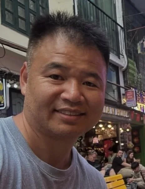 Smiling man with short black hair in an urban street setting with shops and people in the background.