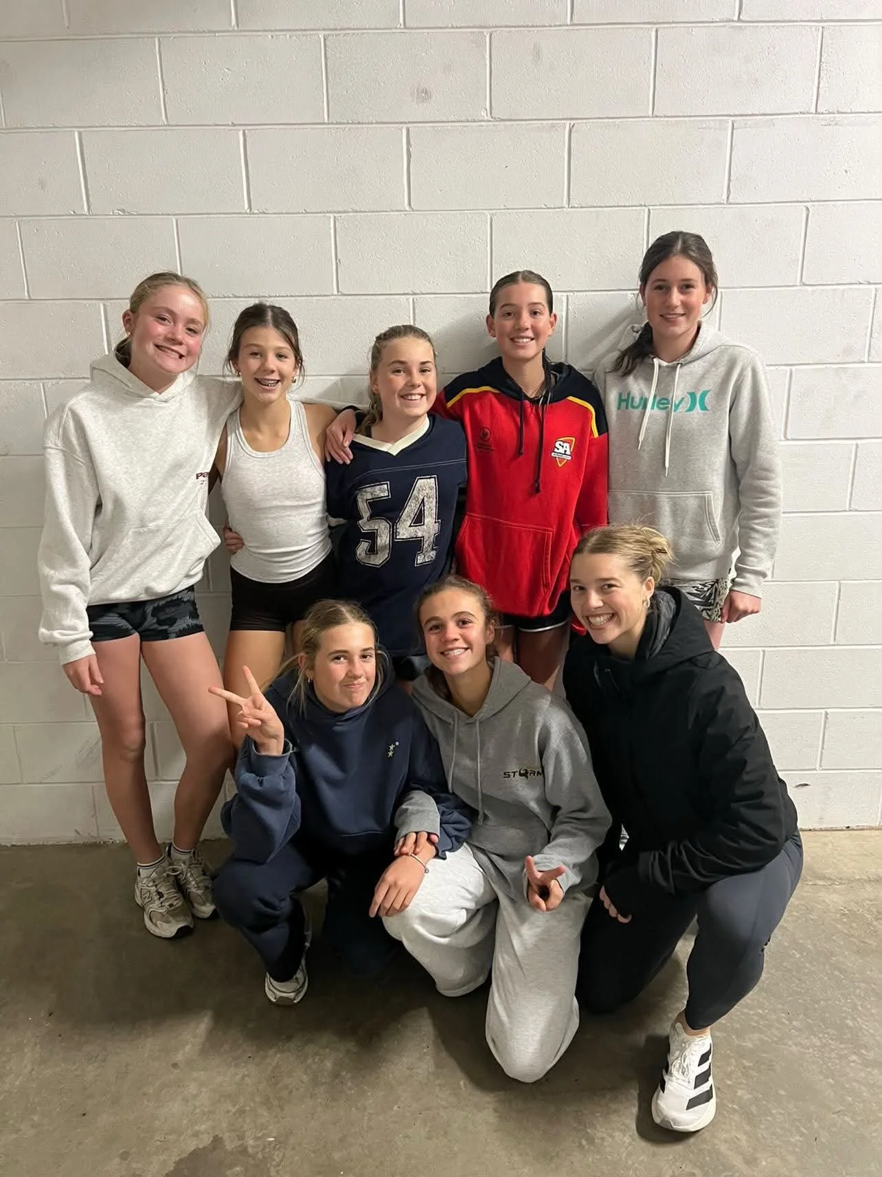 Group of eight young women posing together against a white brick wall, some standing and some kneeling, all smiling and showing camaraderie.