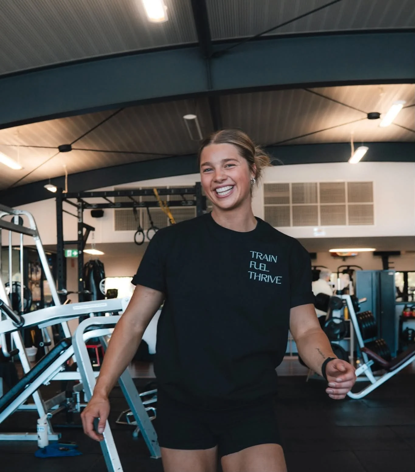 A woman smiling and exercising in a gym, wearing a black t-shirt with the words "Train Fuel Thrive" printed on it.