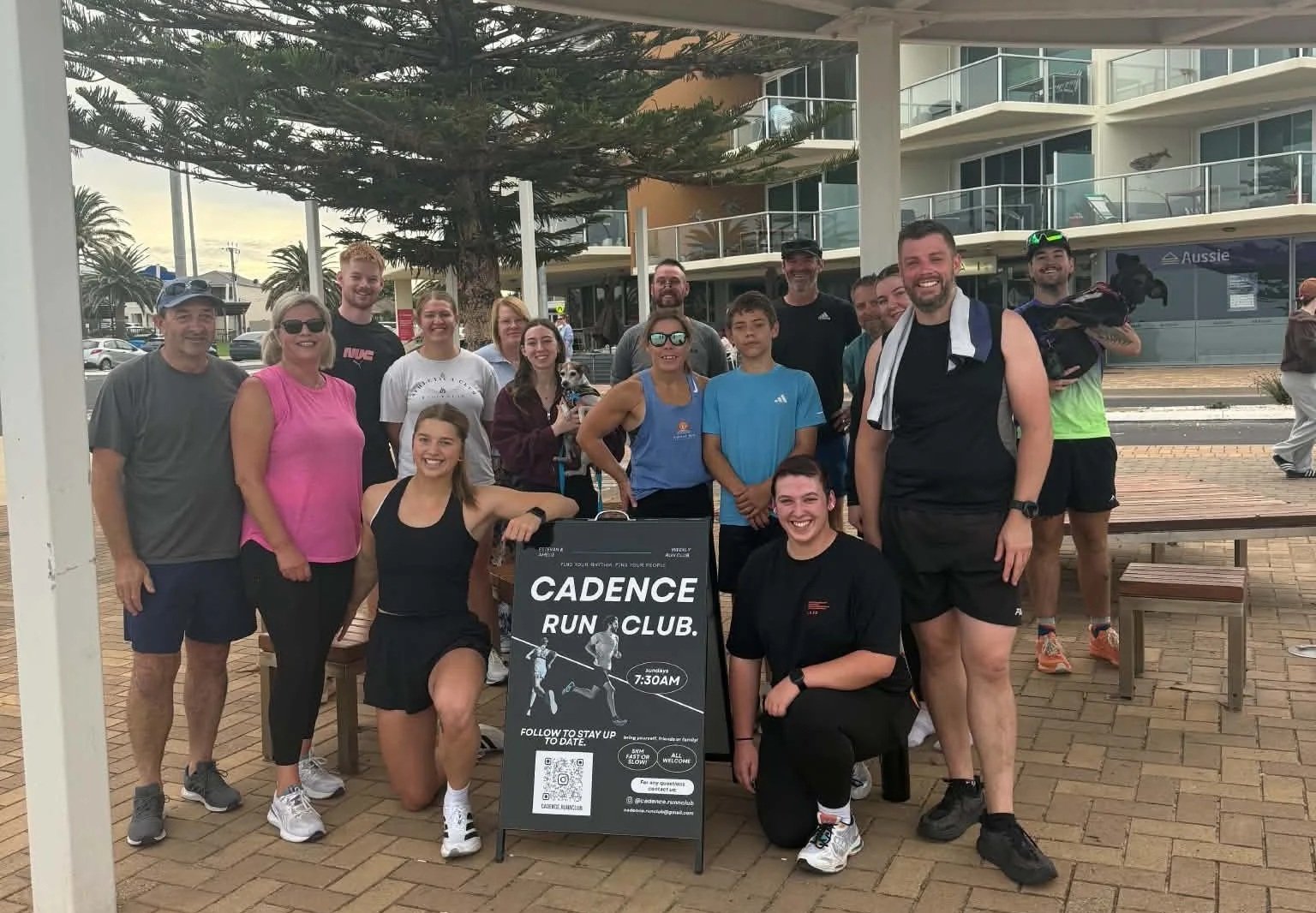 Group of people gathered outdoors around a sign for Cadence Run Club, some smiling and dressed in athletic clothing, with trees and apartment buildings in the background.