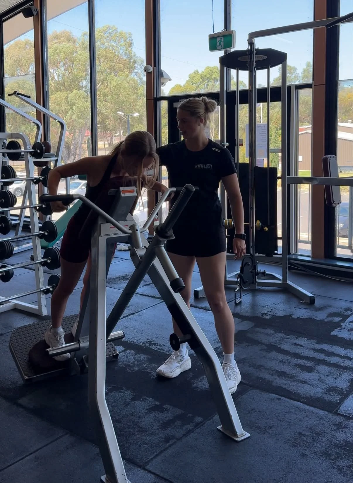 Two women in workout attire in a gym. One is on a step machine, and the other is assisting her. The gym has large windows with trees outside, and various gym equipment is visible.