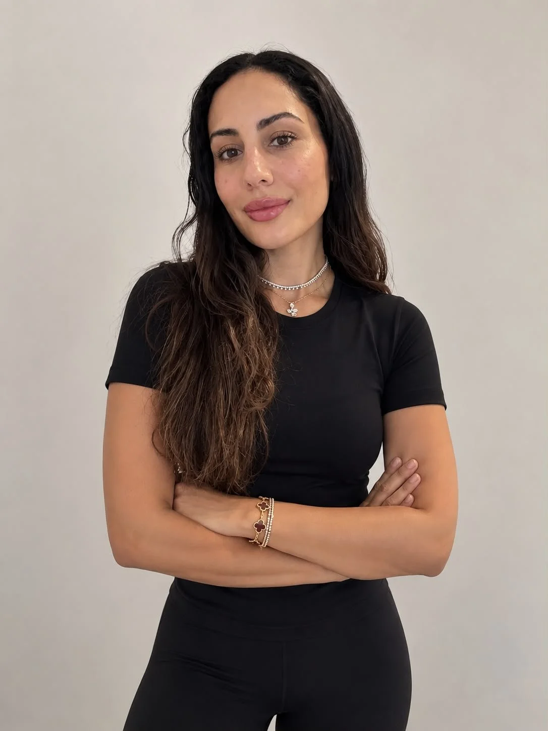 A woman with long dark brown wavy hair standing against a plain light-colored background, wearing a black t-shirt, black leggings, and layered silver necklaces, with her arms crossed, smiling softly at the camera.