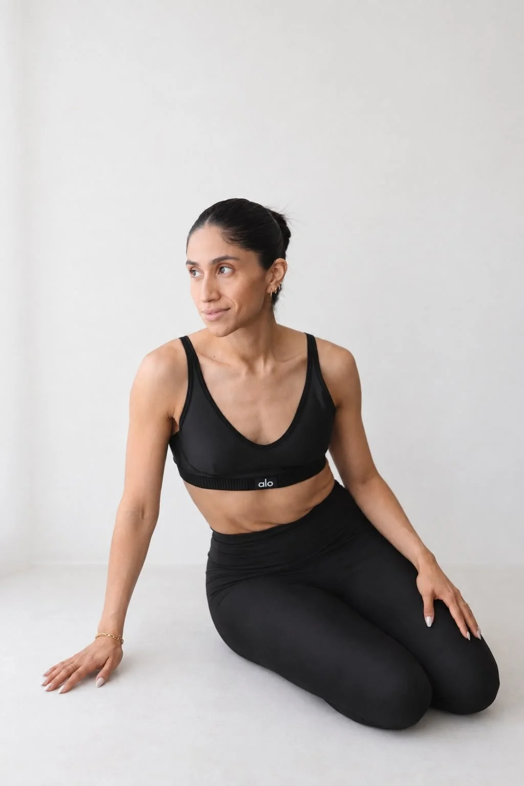 Woman practicing yoga on a white floor against a plain white wall, wearing a black sports bra and black leggings.