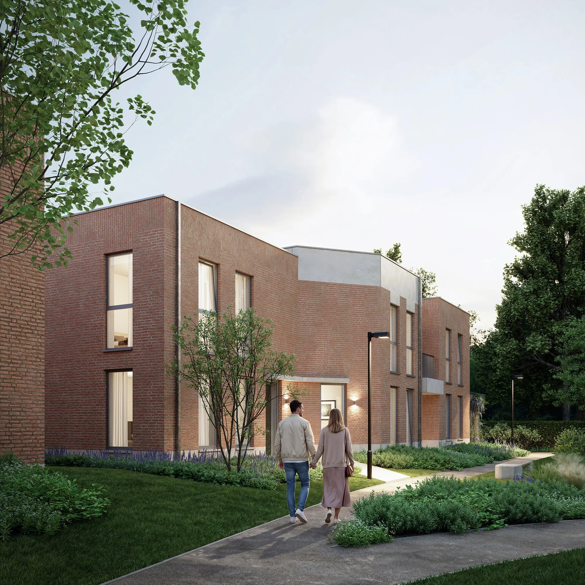 Twee mensen wandelen op een pad naast een modern, bakstenen gebouw met grote ramen, omgeven door een tuin met groene struiken en bomen, 's avonds met buitenverlichting aan.