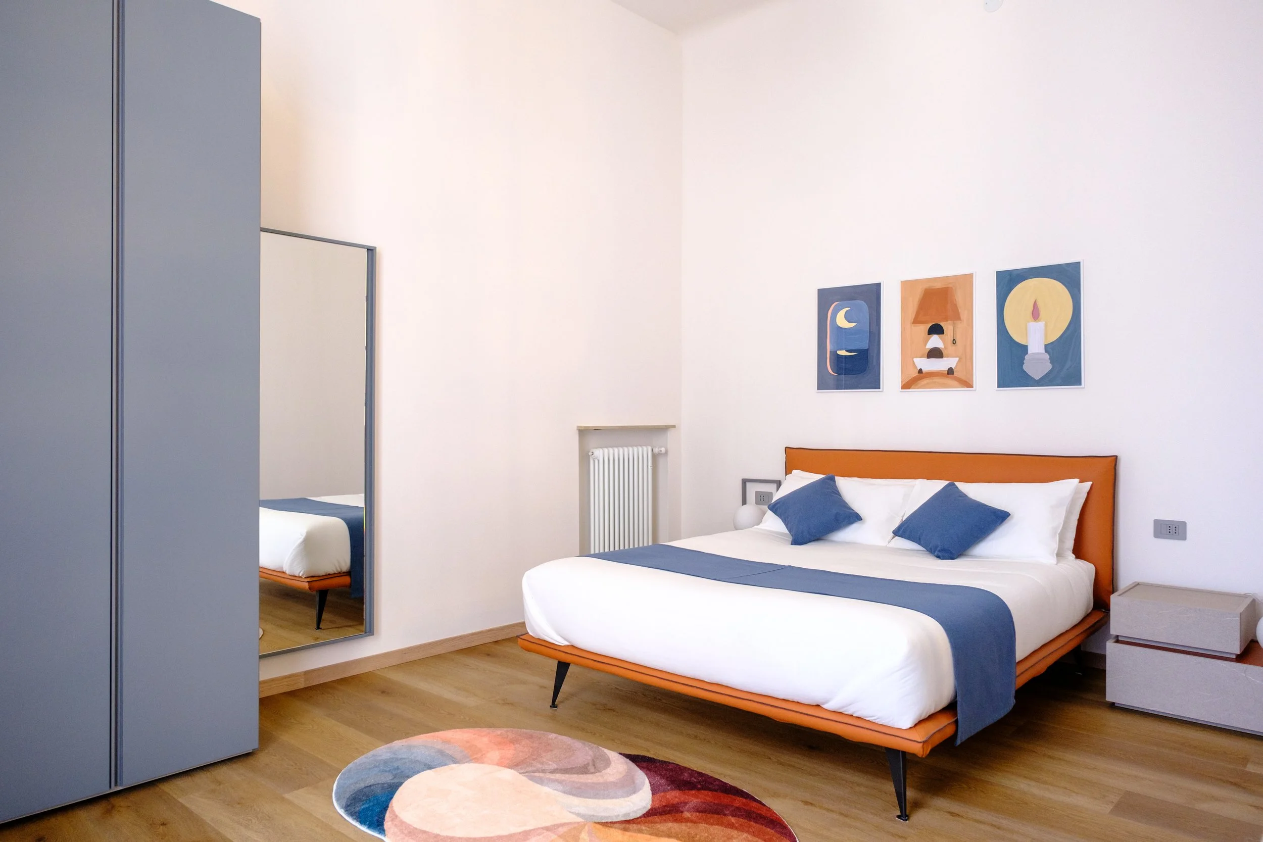 Camera da letto moderna con letto matrimoniale, armadio blu, specchio a parete, tappeto colorato, quadri astratti e arredi in tonalità calde e neutre.
