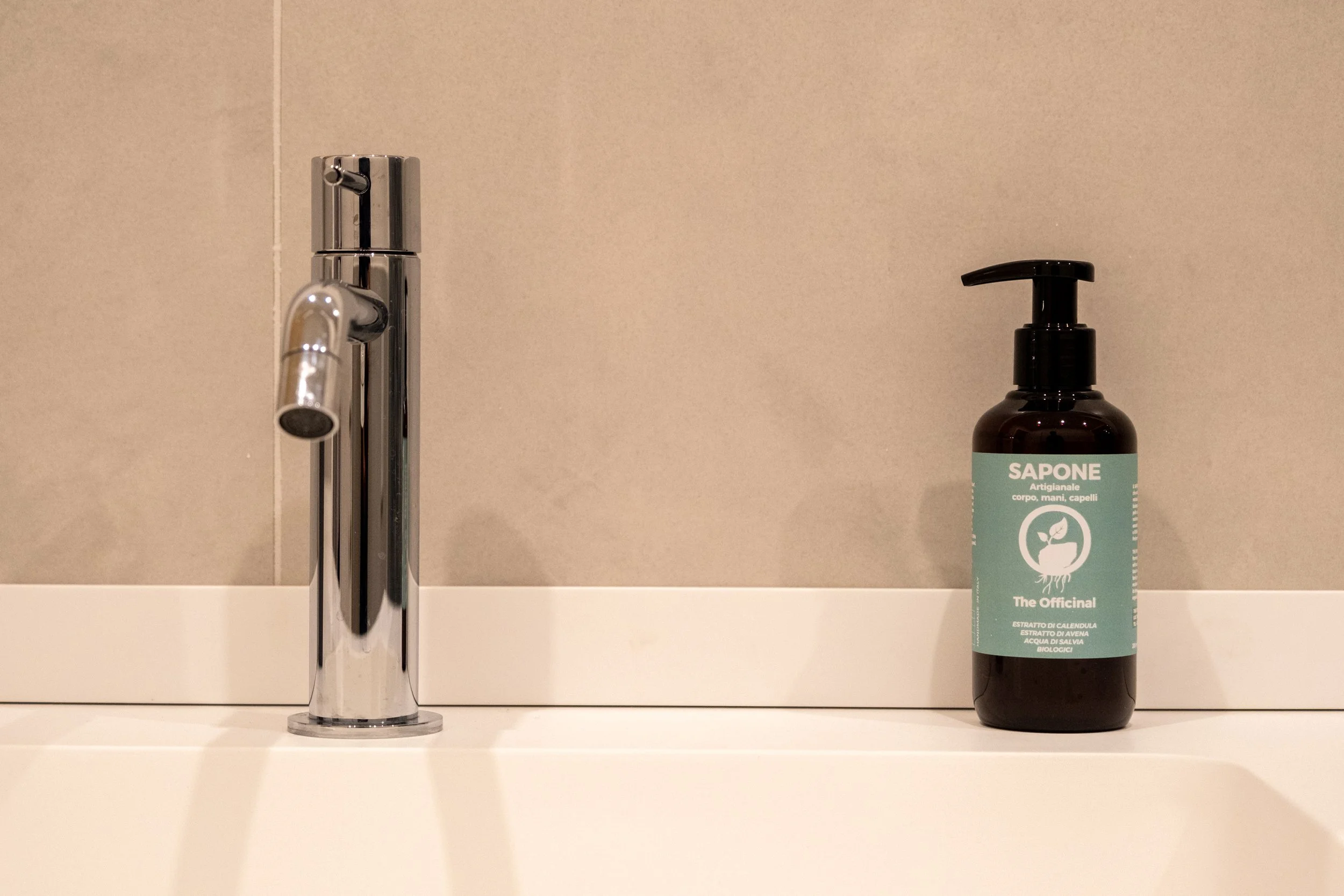 Bottiglia di sapone artigianale con etichetta verde, di fronte a un rubinetto in bagno.