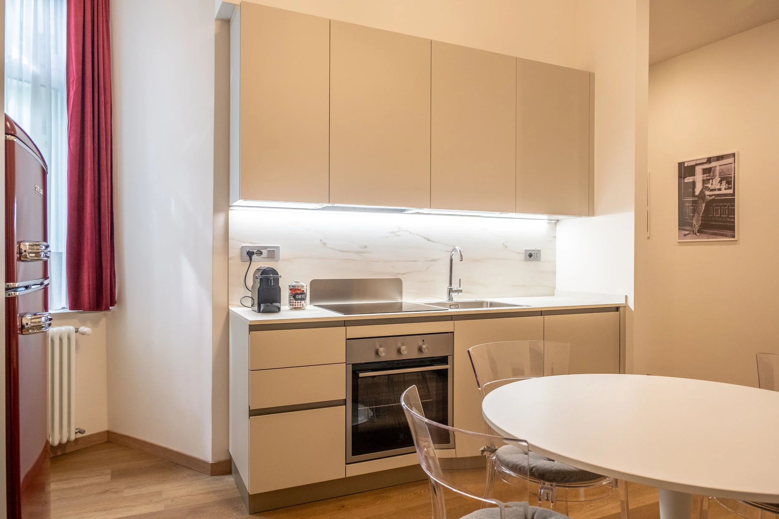 Cucina moderna con mobili beige, forno elettrico, lavello, caffettiera, frigo rosso, tavolo rotondo con sedie trasparenti, pareti chiare e lampada a striscia sotto gli armadi.