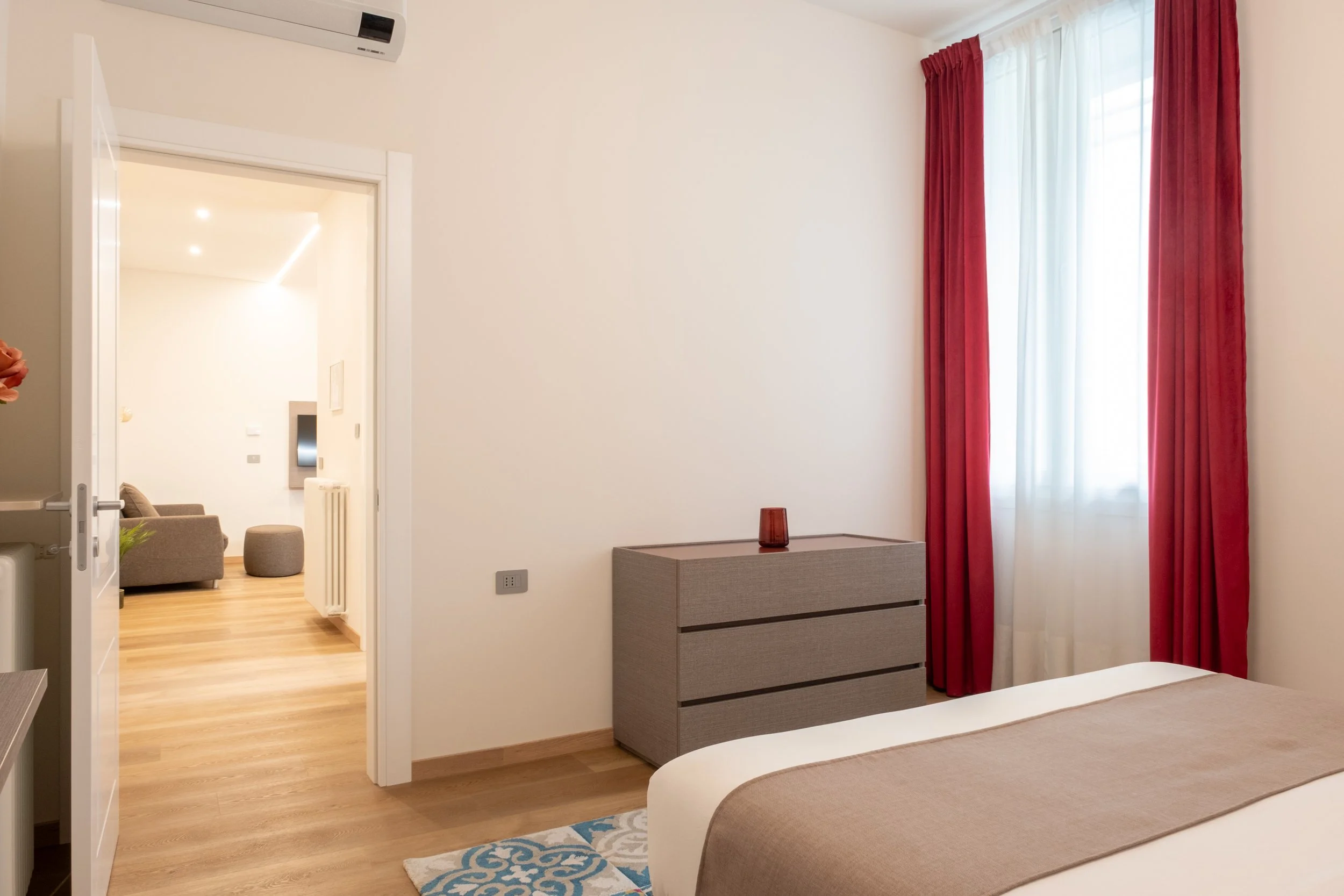 Camera da letto con finestra con tende rosse, comò grigio, letto con coperta beige, diposizione moderna, ingresso a un soggiorno con divano e tavolino, pavimento in legno.