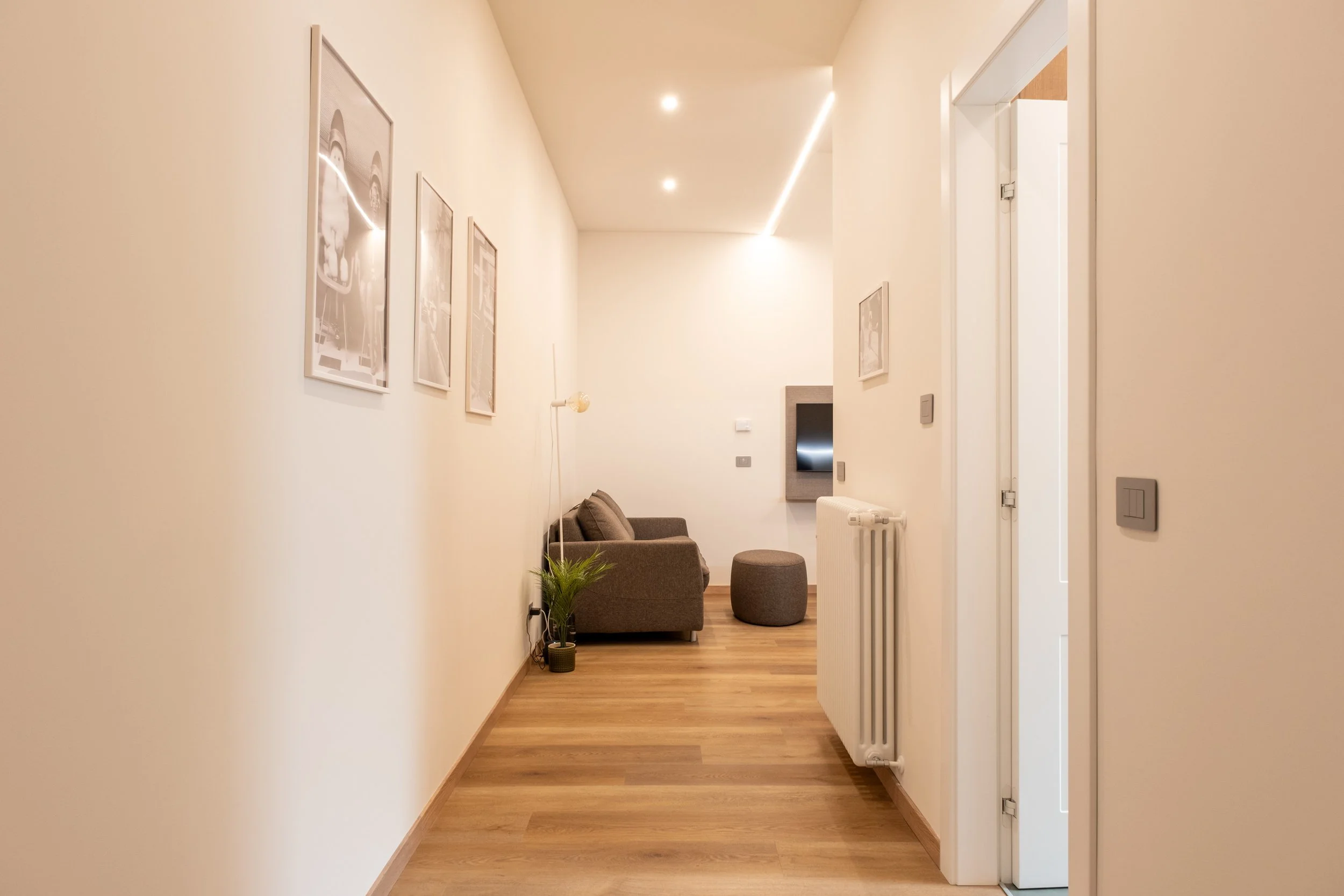 Render di un'ampia sala soggiorno con pareti beige, pavimento in legno e arredi minimalisti, tra cui un divano marrone, un tavolino e una tv appesa alla parete.