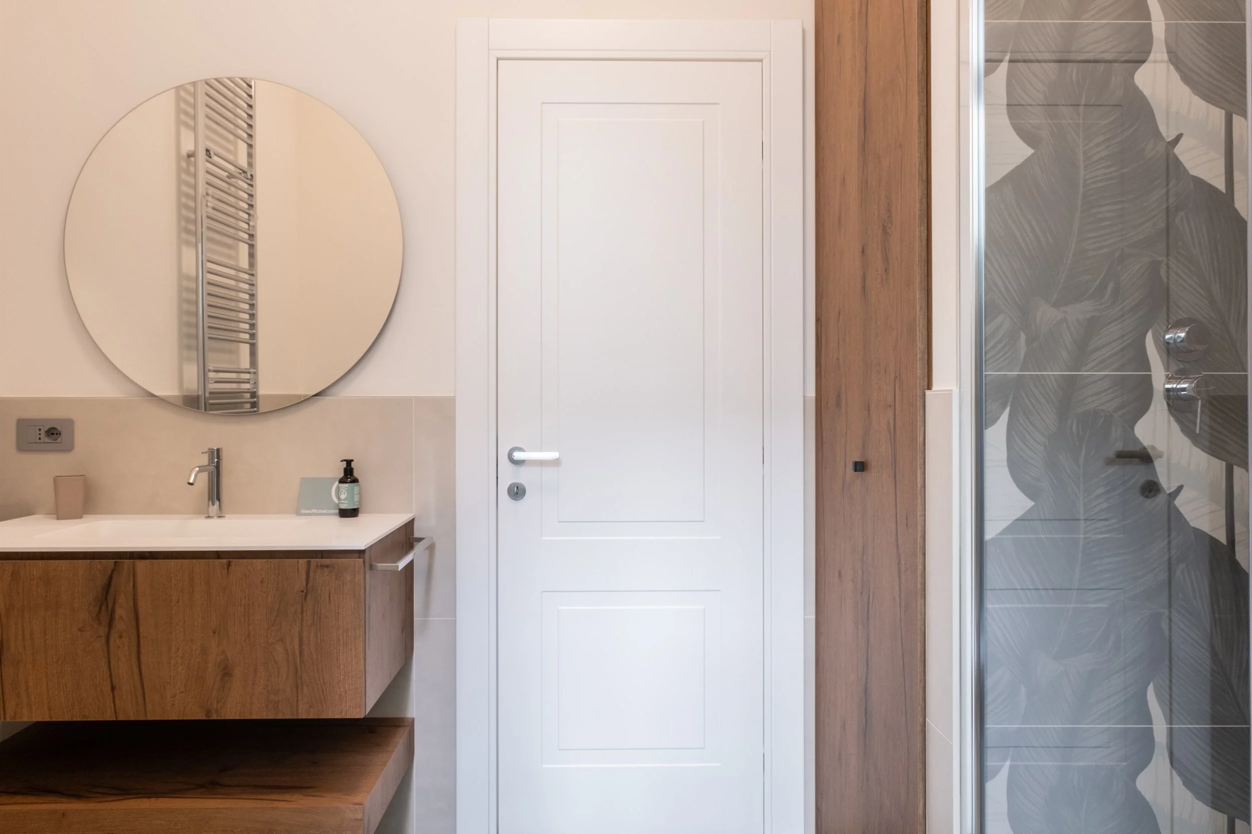 Bagno con mobile in legno, grande specchio tondo e cabina doccia con pareti decorate con foglie