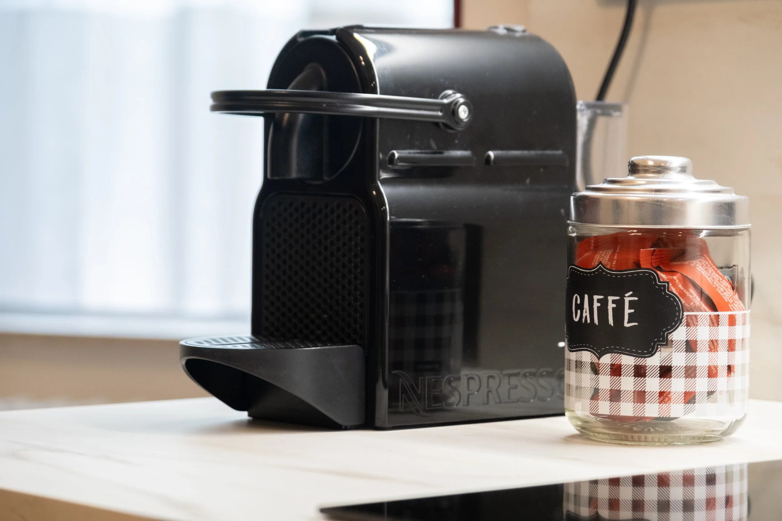 Caffettiera Nespresso nera accanto a un barattolo di vetro con etichetta 'Caffé' e contenuto di bustine di zucchero, su un tavolo di legno.