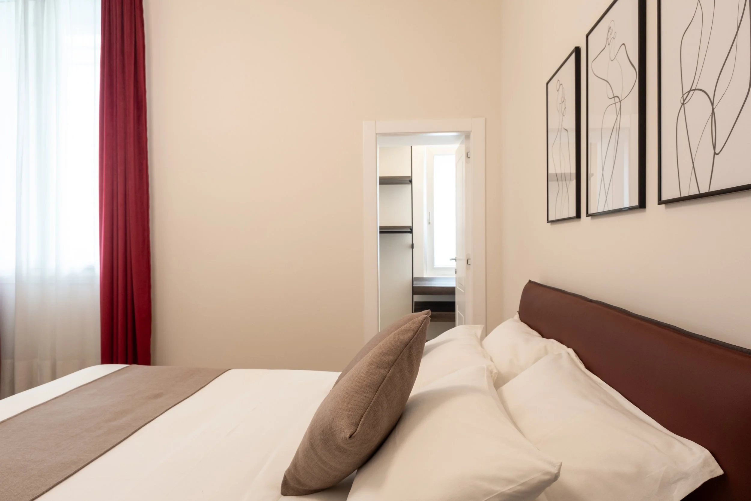 Camera da letto con letto con lenzuola bianche, cuscini beige, pareti beige, quadri con linee artistiche, finestra con tende rosse e bianche e porta aperta che porta a un piccolo spogliatoio.