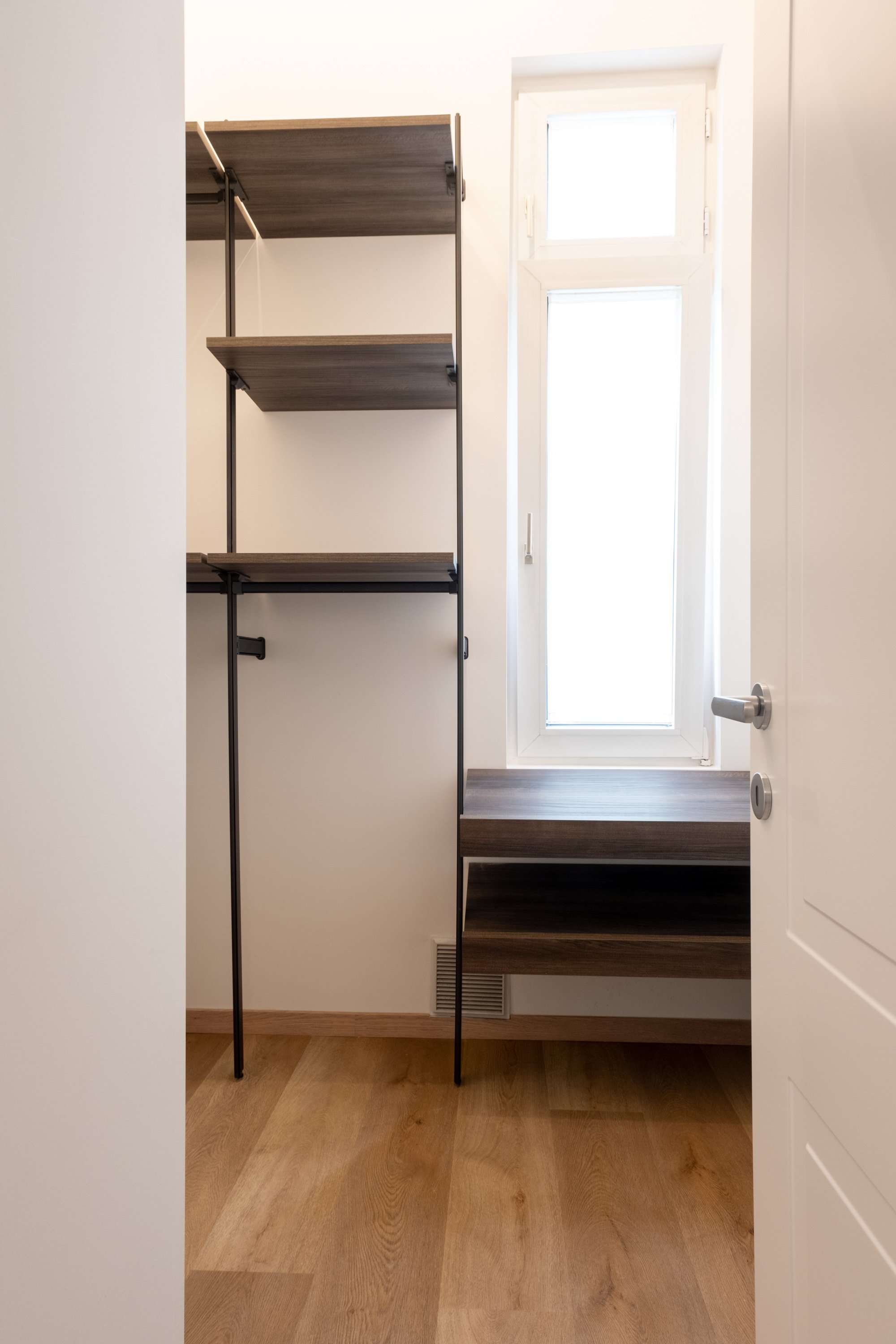 Stanza con scaffale a parete in metallo e pannelli di legno, finestra grande con vetri trasparenti, pavimento in legno chiaro.