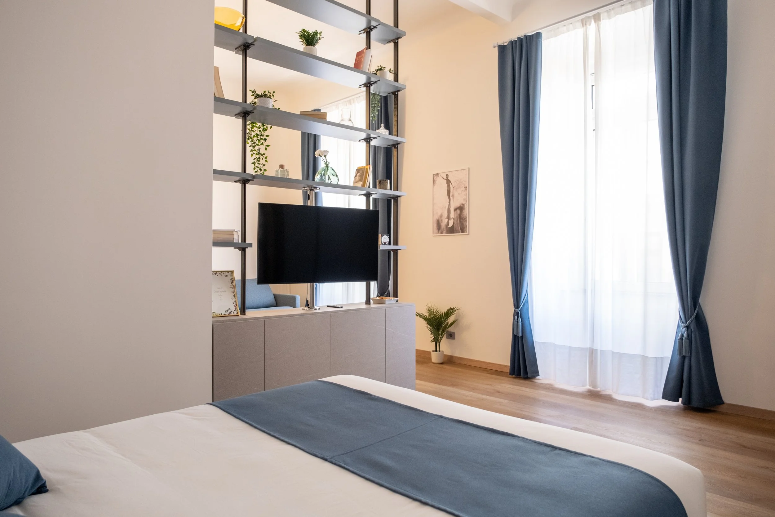 Camera da letto moderna con letto con copriletto blu, tende blu e pareti chiare, scaffali aperti con piante e decorazioni, televisore, e finestra con tendaggi lunghi.