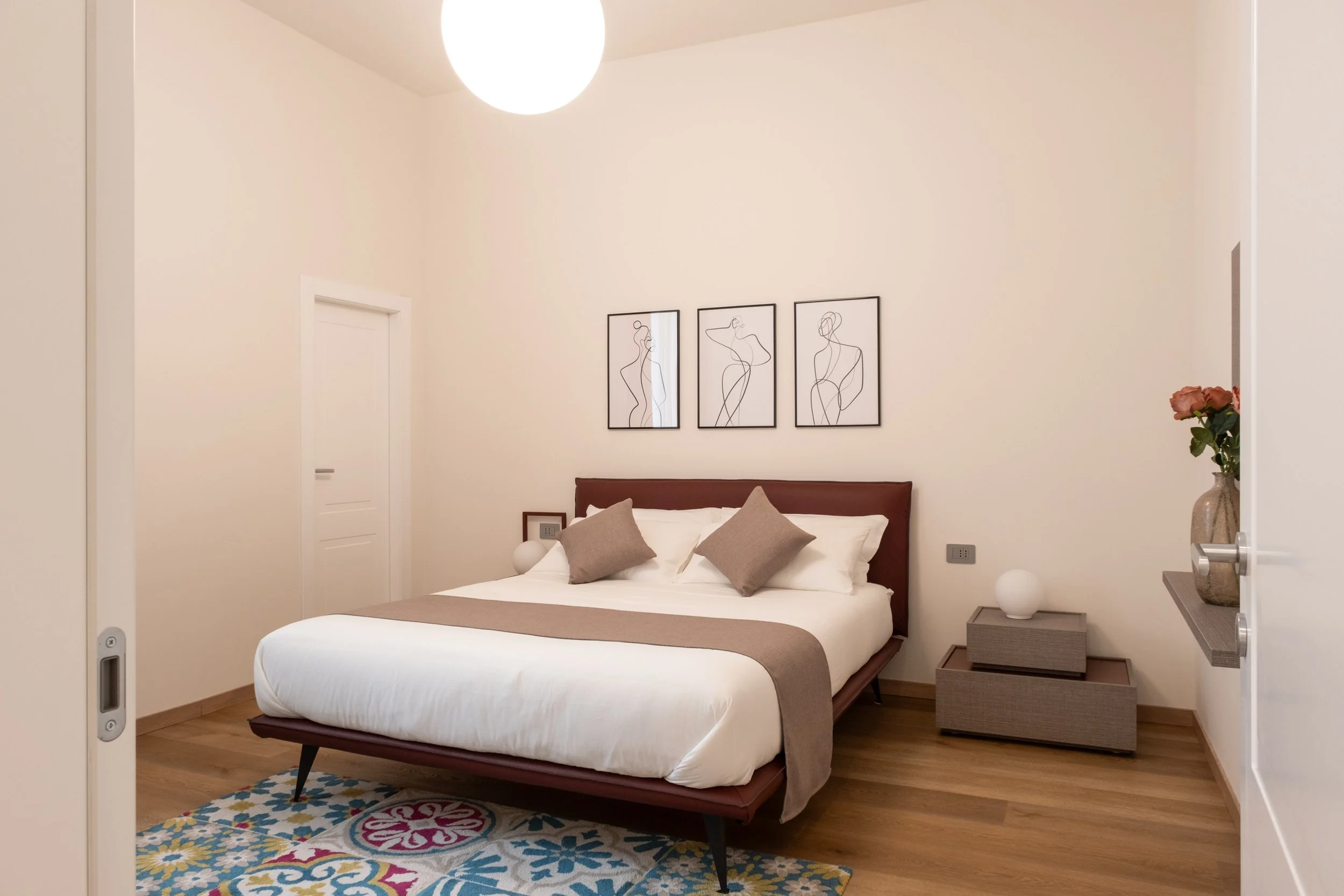 Camera da letto moderna con letto matrimoniale con testiera in pelle marrone, quattro cuscini beige, quadri astratti in bianco e nero sopra il letto, due comodini con lampade bianche, tappeto colorato e pianta con fiori arancioni.