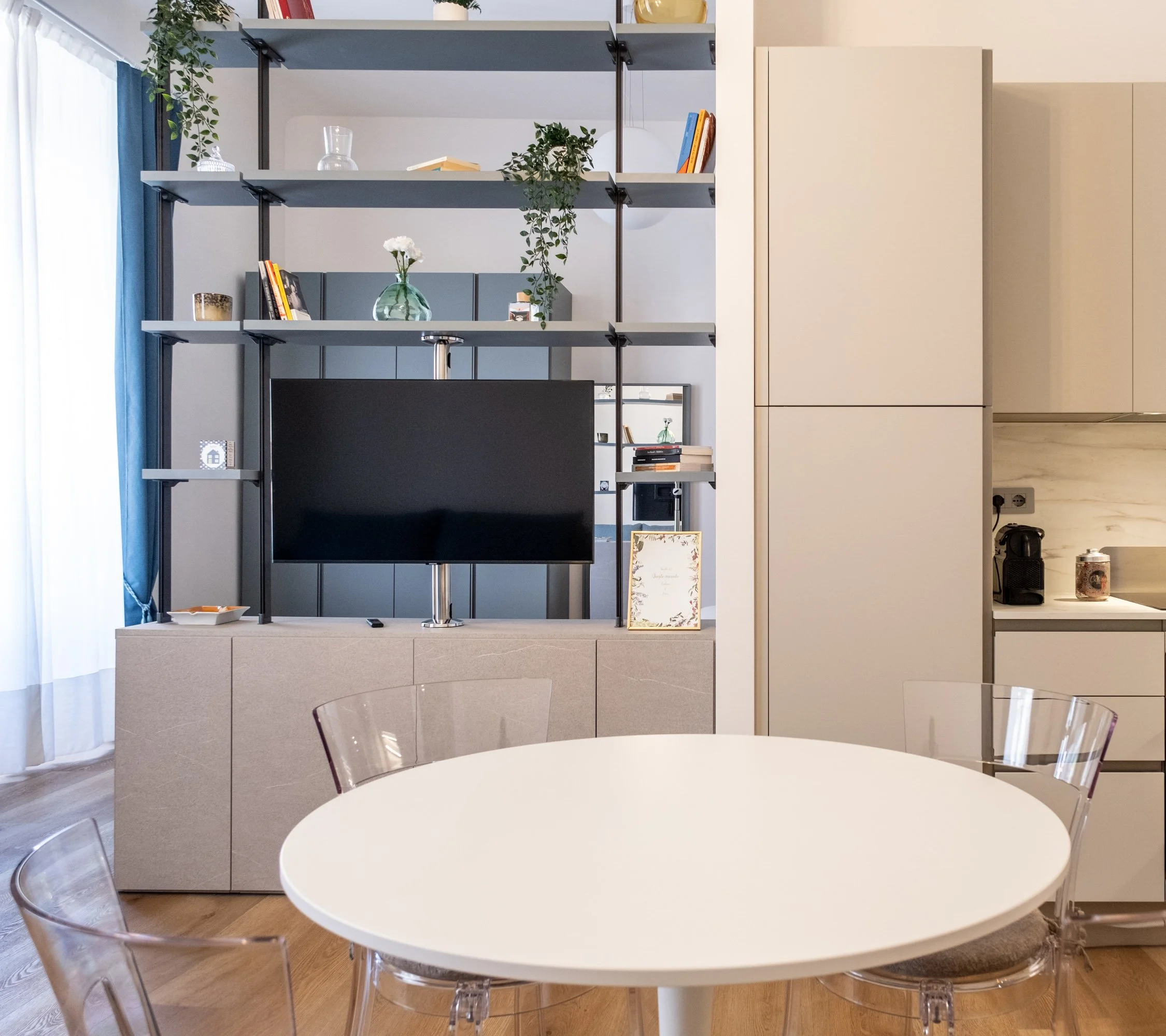 Interno di una zona living con tavolo rotondo bianco e sedie trasparenti, libreria moderna con television, piante e libri, e cucina in background.
