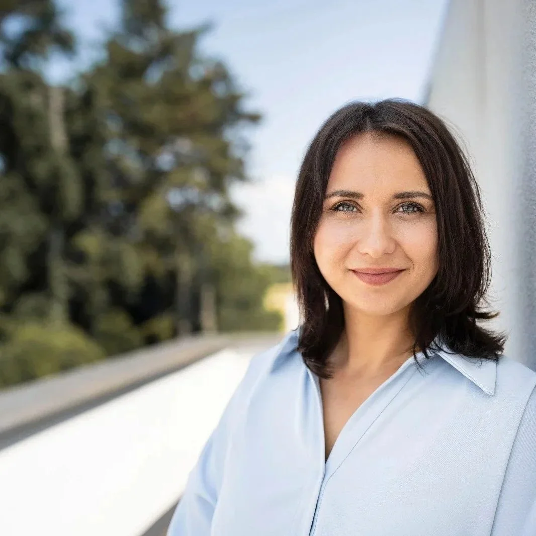 Nahaufnahme einer lächelnden Frau mit braunen, schulterlangen Haaren, in einem weißen Hemd, steht draußen an einer Wand, im Hintergrund sind Bäume und ein blauer Himmel sichtbar.