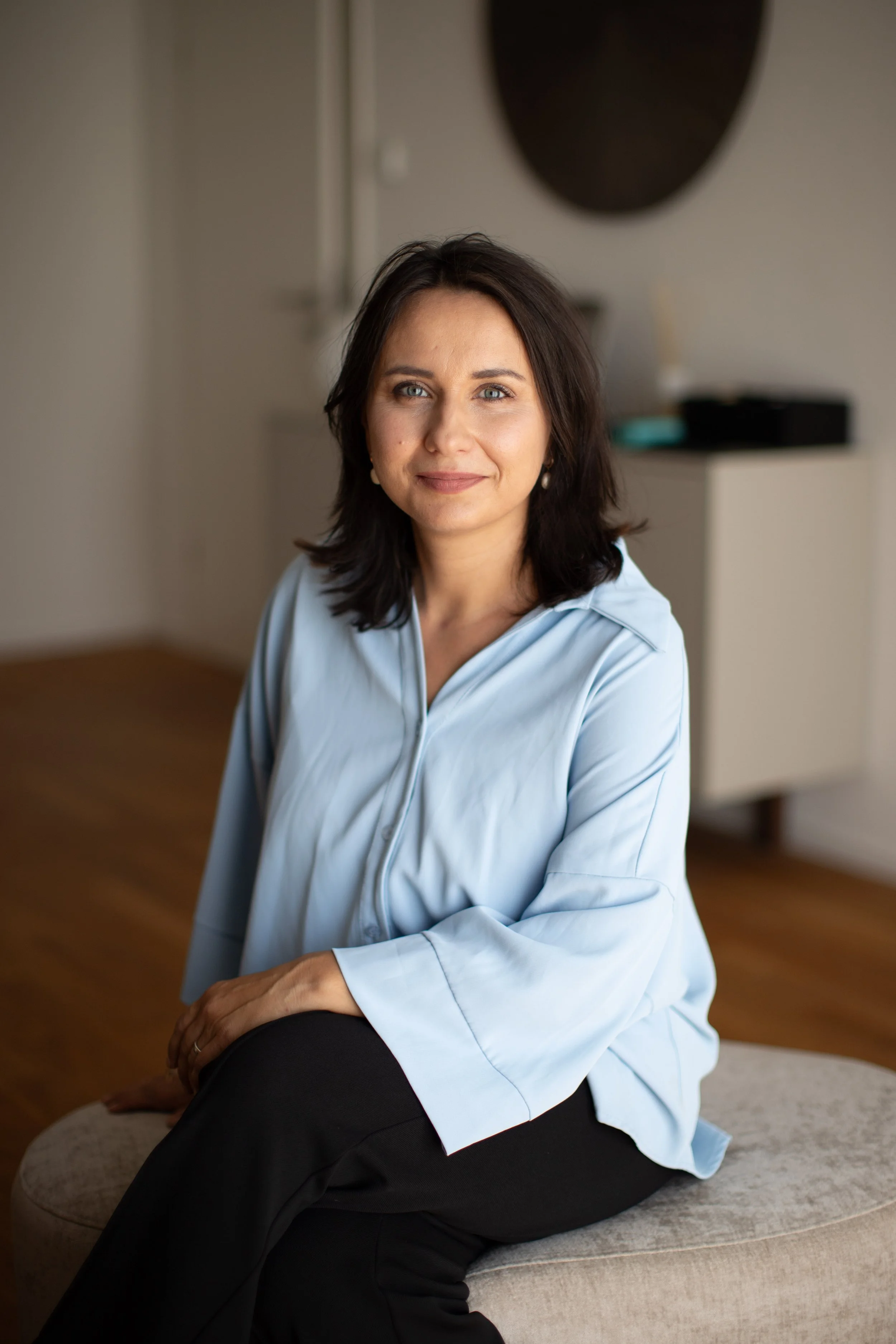 Eine Frau mit dunklen, schulterlangen Haaren, trägt eine hellblaue Bluse und sitzt auf einem Hocker in einem modernen, hell eingerichteten Beratungsraum.