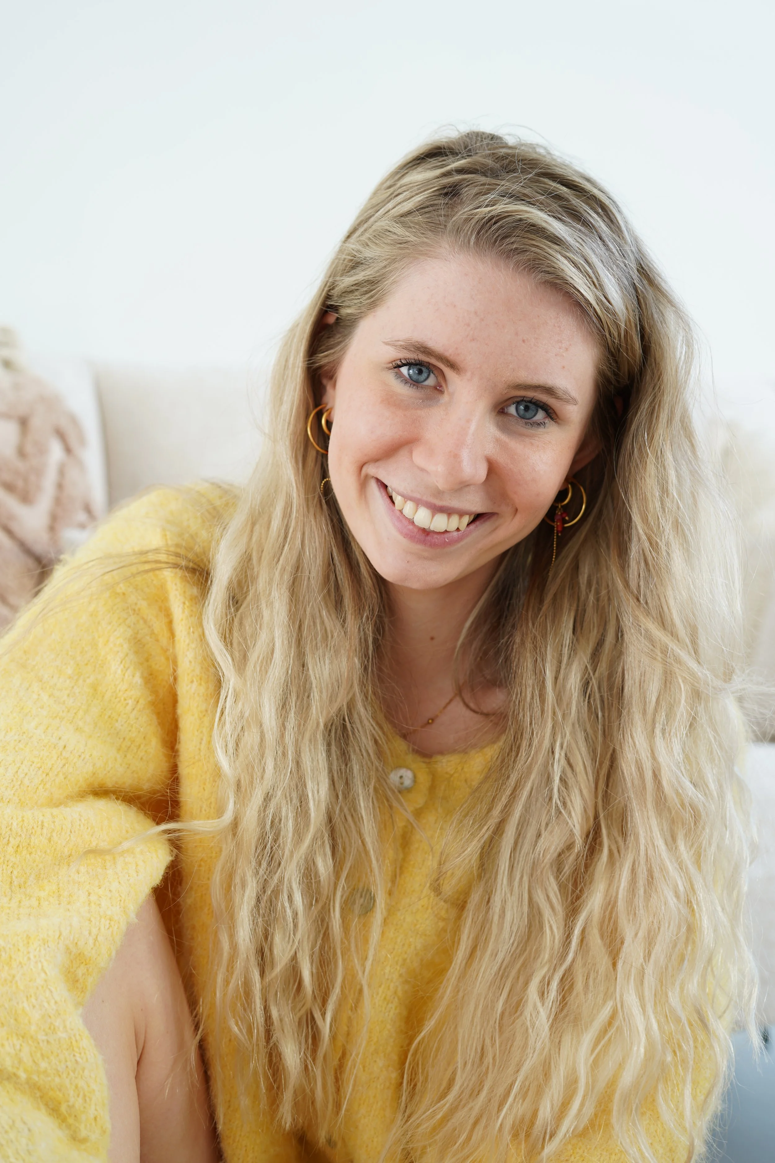 A smiling young woman with long blond wavy hair wearing a yellow cardigan and gold hoop earrings.