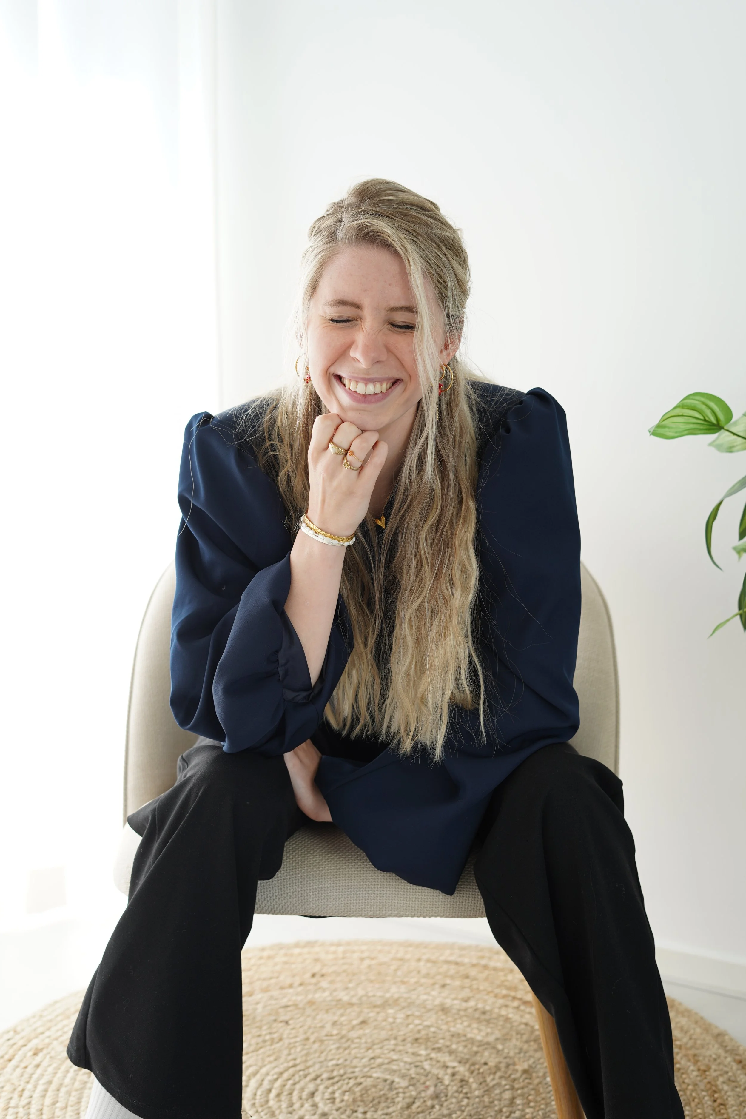A woman with long blonde hair, wearing a navy blue blouse and black pants, is sitting on a beige chair, smiling with her eyes closed and her right hand resting on her chin. There is a green plant to her right and a woven rug on the floor.