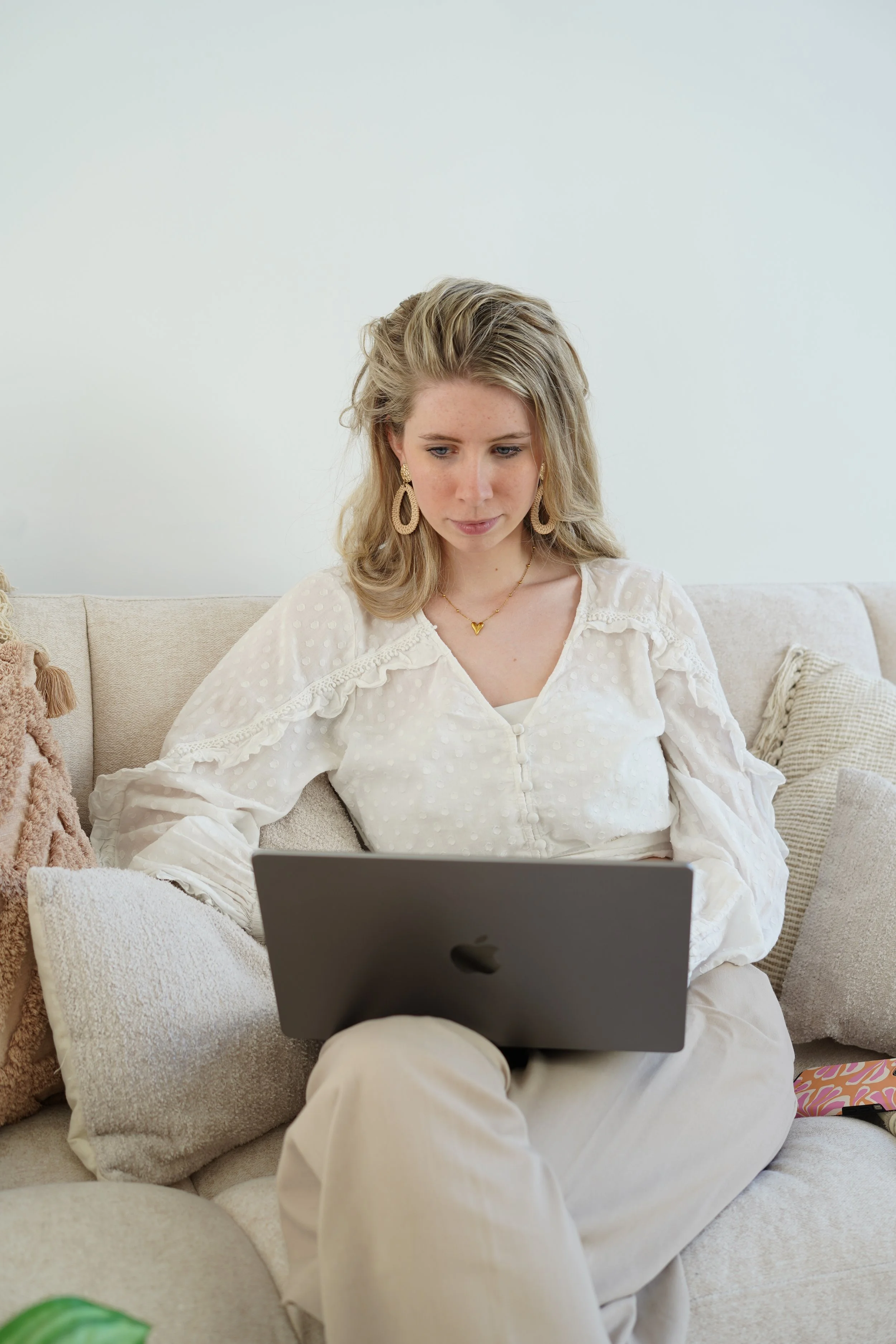 A woman with blonde hair, wearing earrings and a white top, sitting on a beige sofa with various cushions, using a laptop.