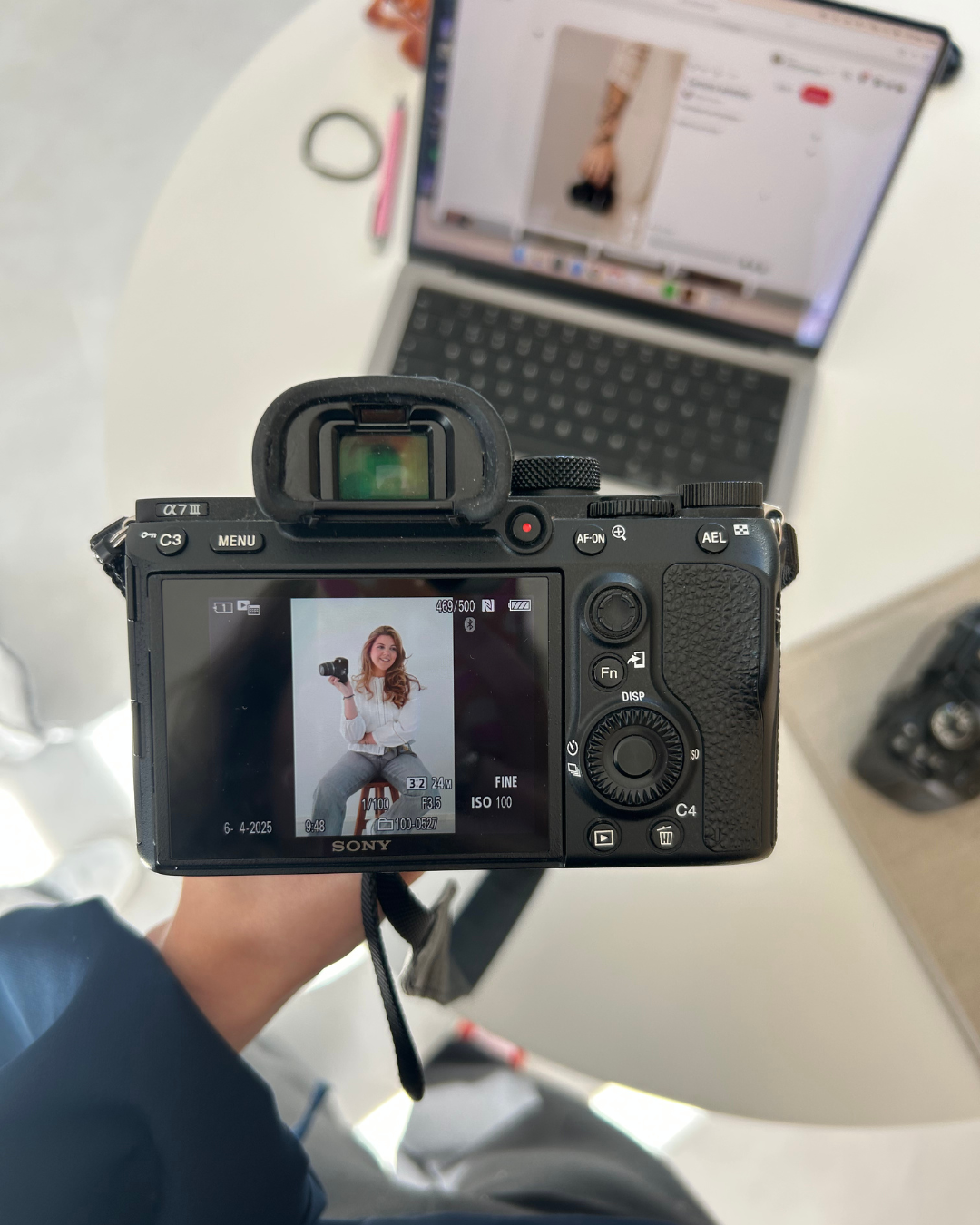 The photo displays a digital camera capturing an image of a woman sitting on a stool, holding a camera, with a laptop in the background on a white table.