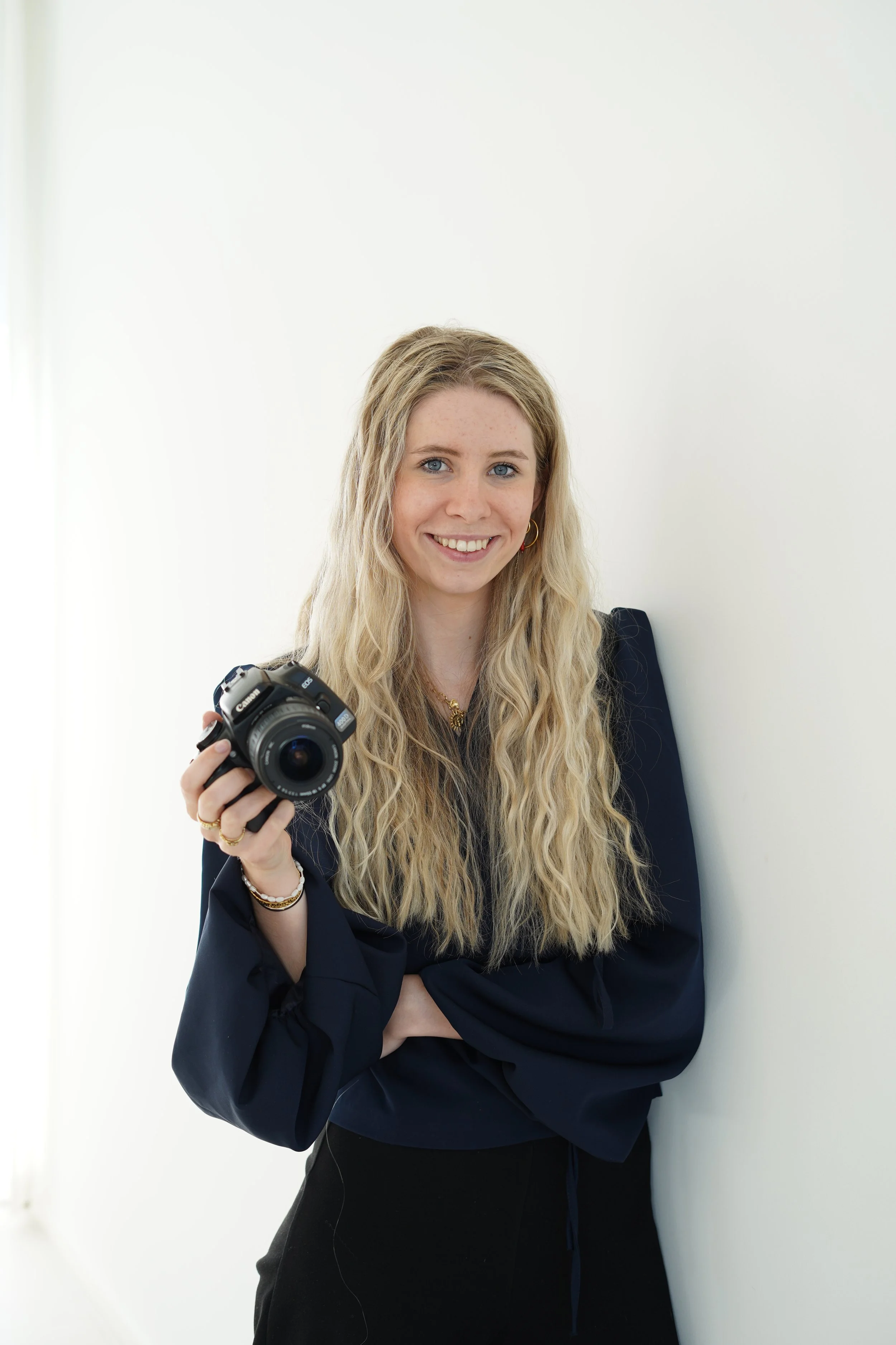 Young woman with long blonde hair, smiling, holding a camera in her right hand, standing against a plain white background.