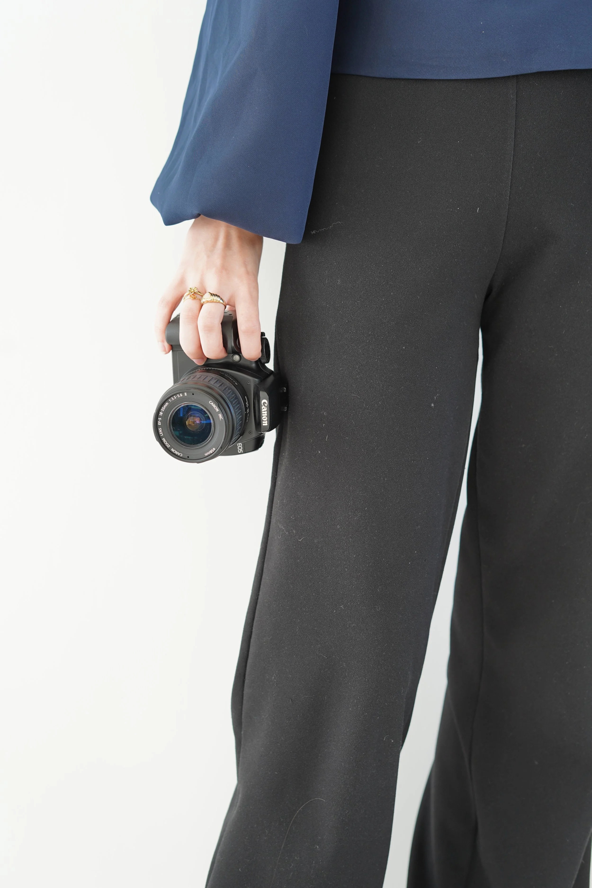 Person wearing black pants and a navy blue blazer, holding a compact black Canon camera with their right hand.