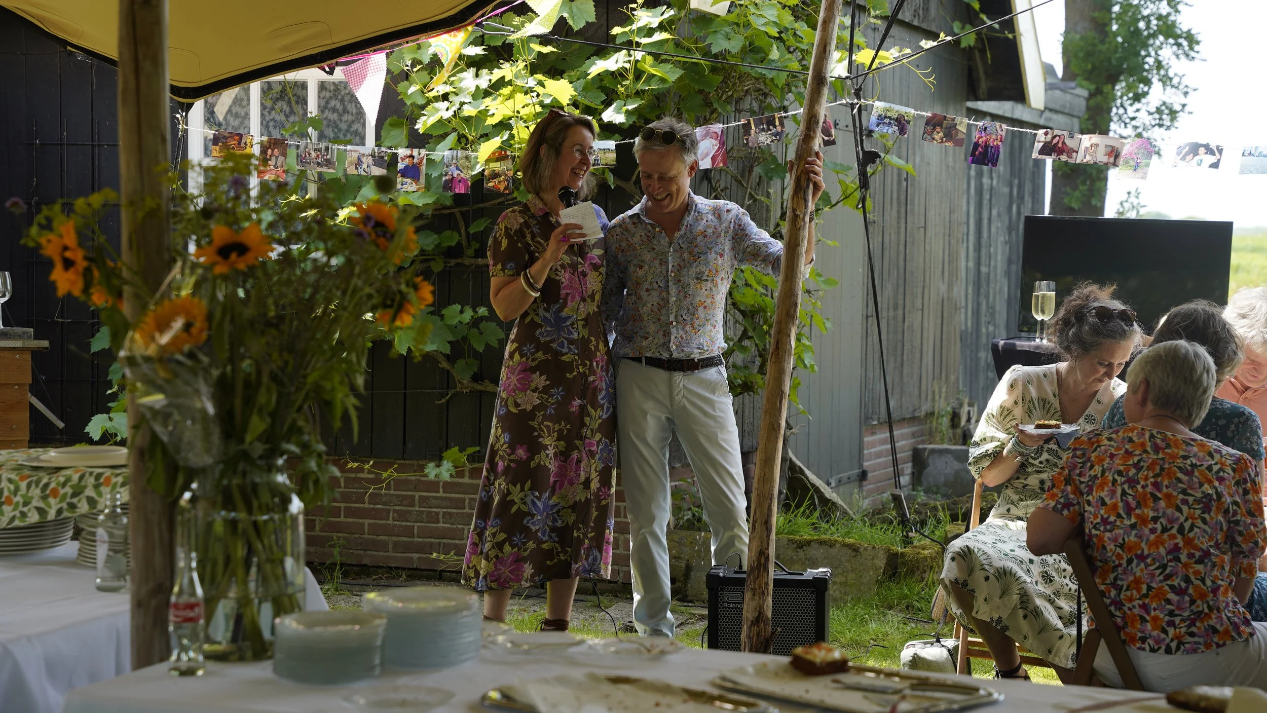 People at a backyard gathering, with two women in floral dresses sharing a moment, one holding a microphone. Guests are seated, some eating, and decorations include photos hanging on a line with greenery in the background.