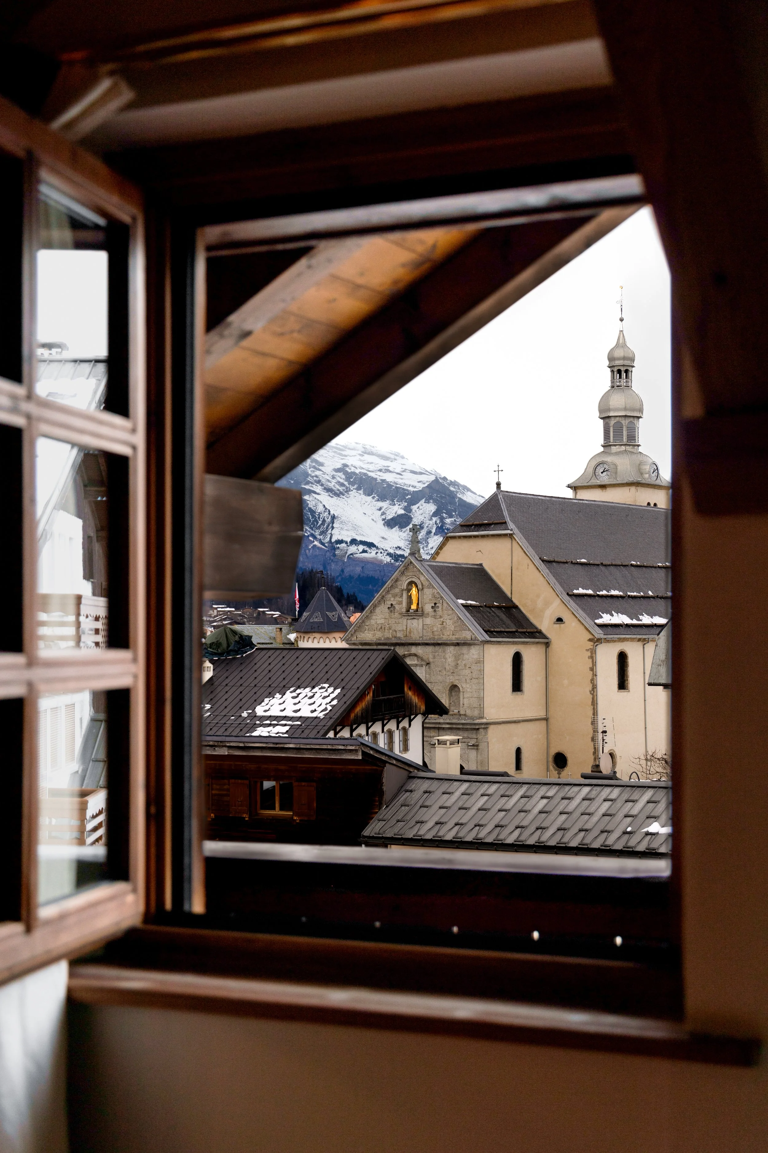 Vue d'une église située dans une ville alpine, vue depuis une fenêtre en bois, avec des montagnes enneigées en arrière-plan.