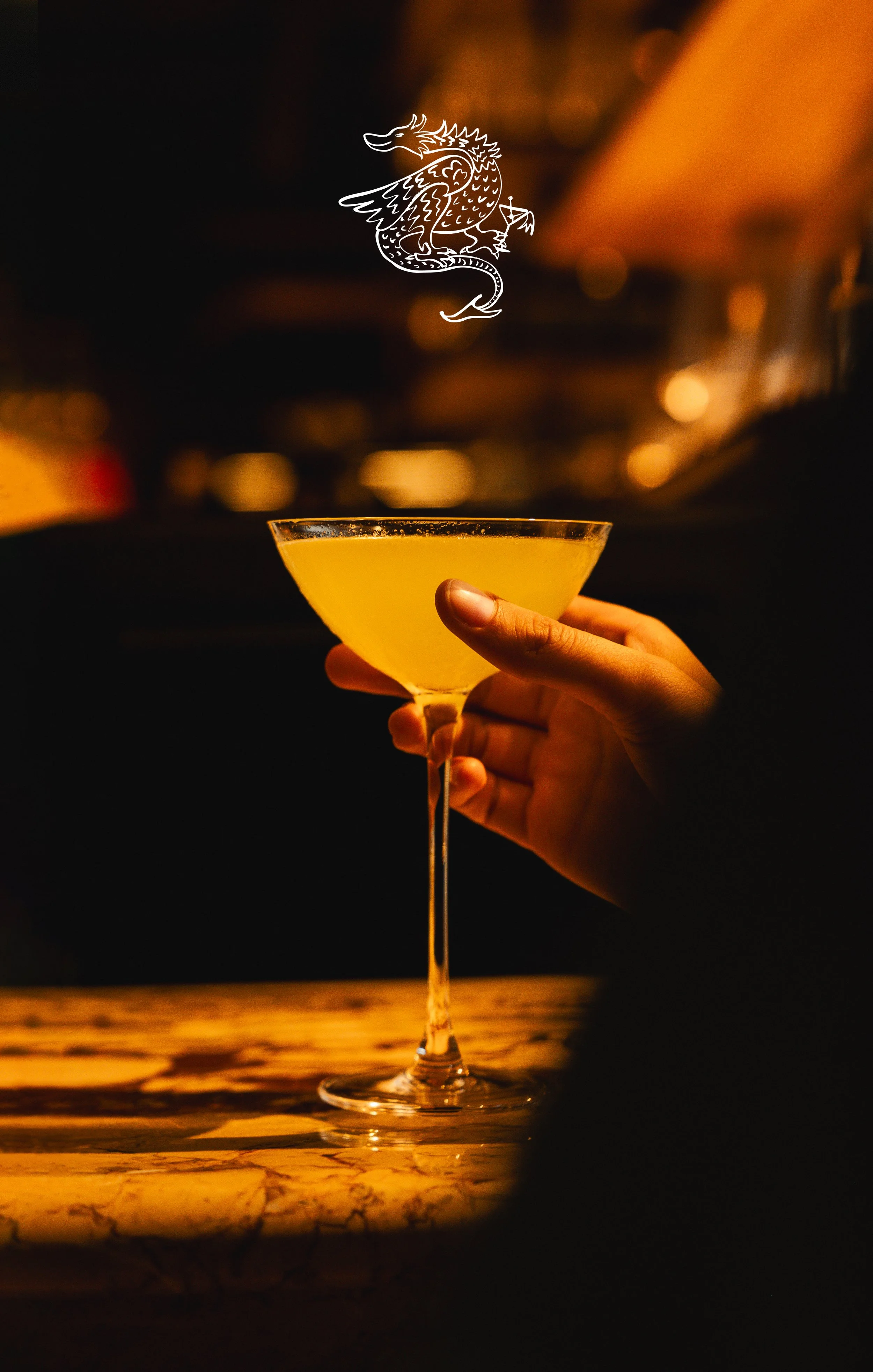 Main tenant un verre à cocktail jaune dans un bar sombre, fond flou avec des lumières chaudes.