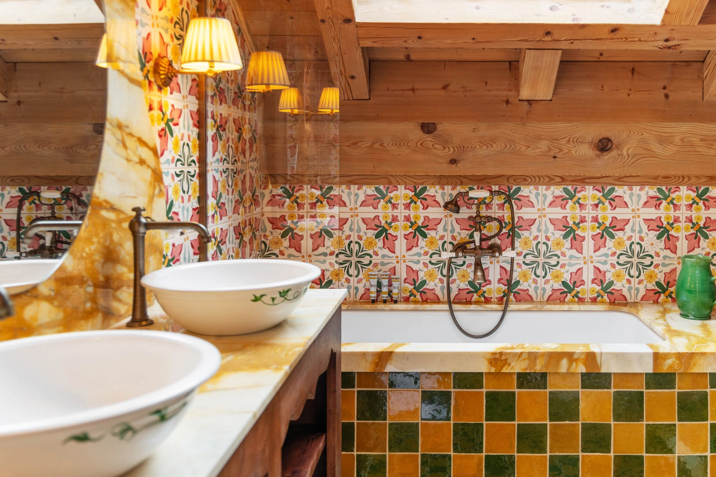 Salle de bain avec murs en carreaux colorés, baignoire encastrée, deux vasques en céramique, éclairage avec appliques murales, plafond en bois.