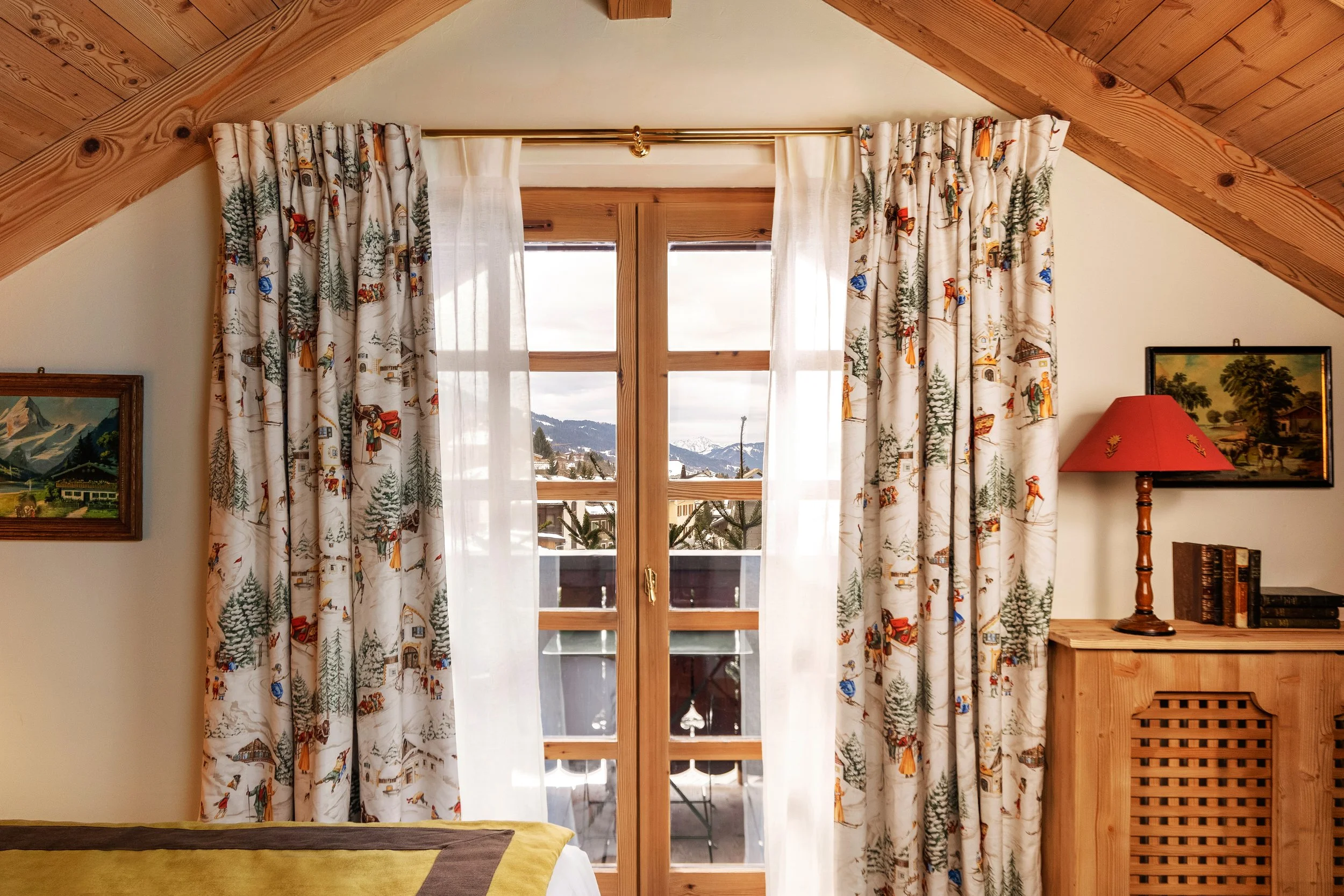 Une vue à travers une porte-fenêtre avec des rideaux à motifs champêtres, donnant sur un balcon avec une vue sur des montagnes enneigées.