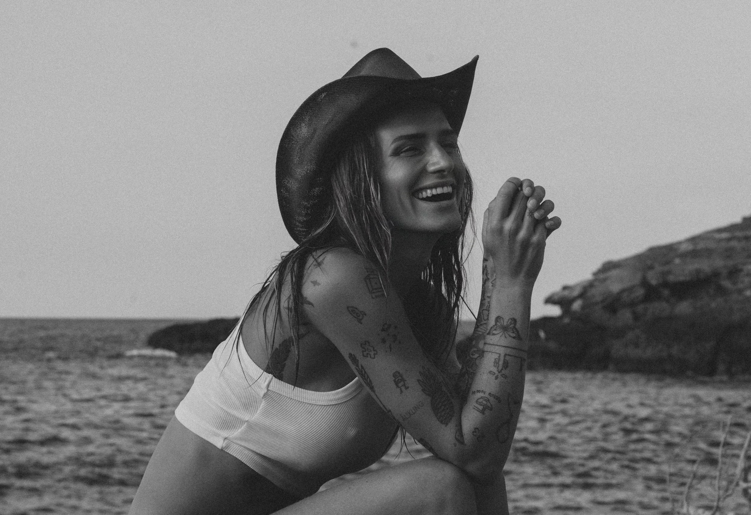Black and white photo of a smiling woman wearing a cowboy hat, sitting outdoors near water with rocks in the background. She has tattoos on her arms and is wearing a sleeveless top.