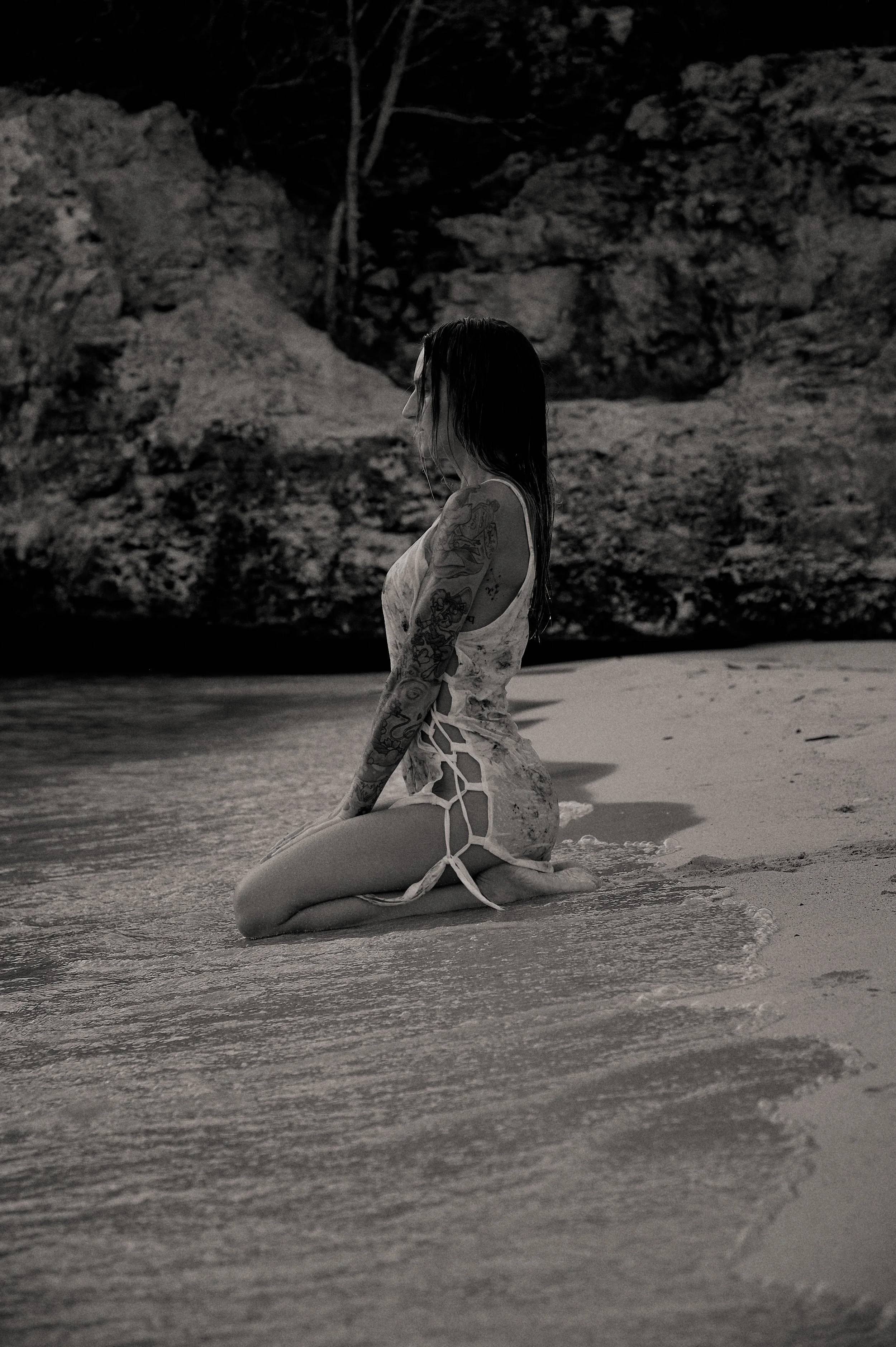 A woman with tattoos kneeling in shallow water on a beach, with rocky cliffs in the background, in black and white.