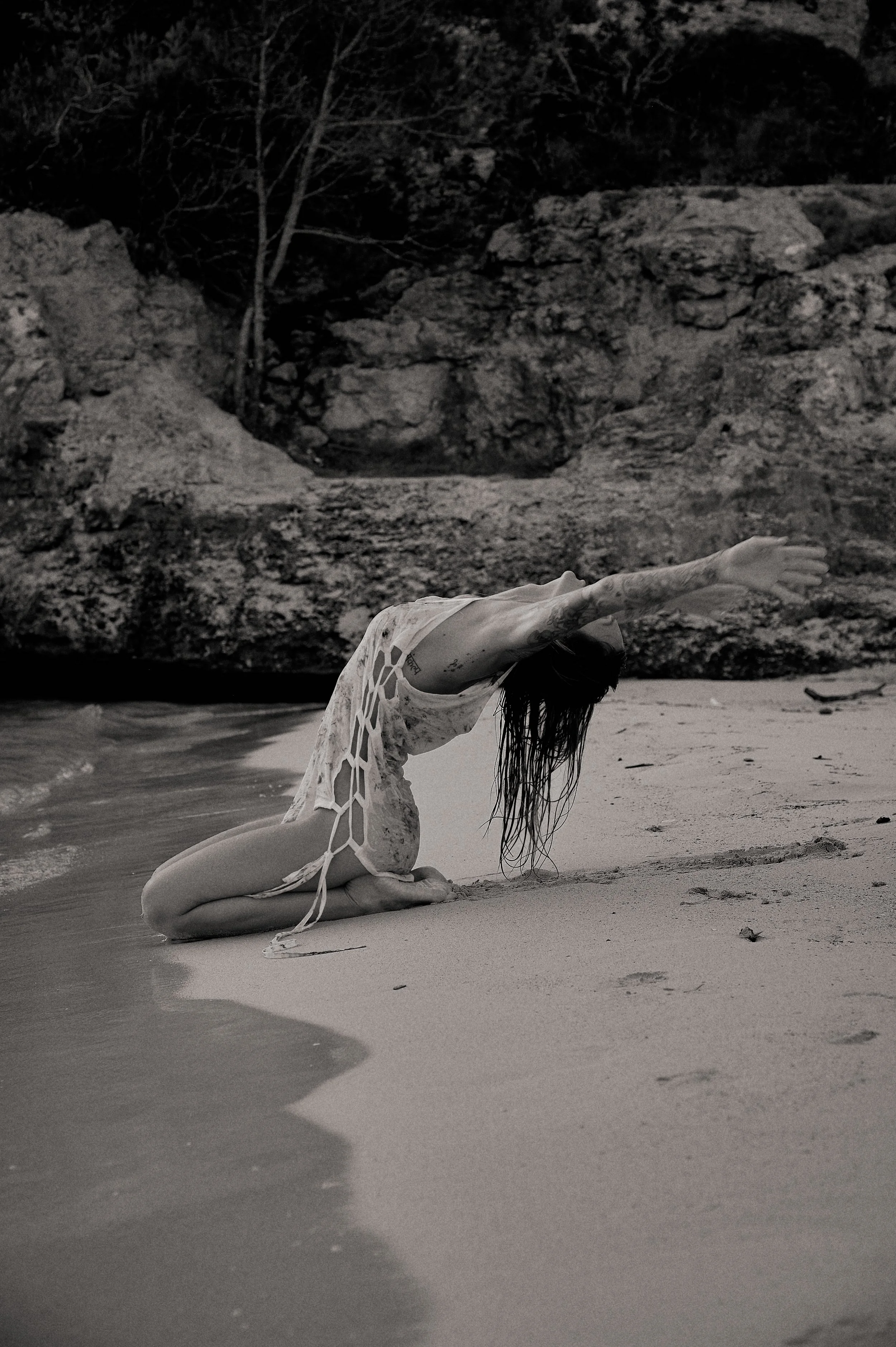 A woman in a lace dress performing a yoga pose on a beach with rocks in the background.