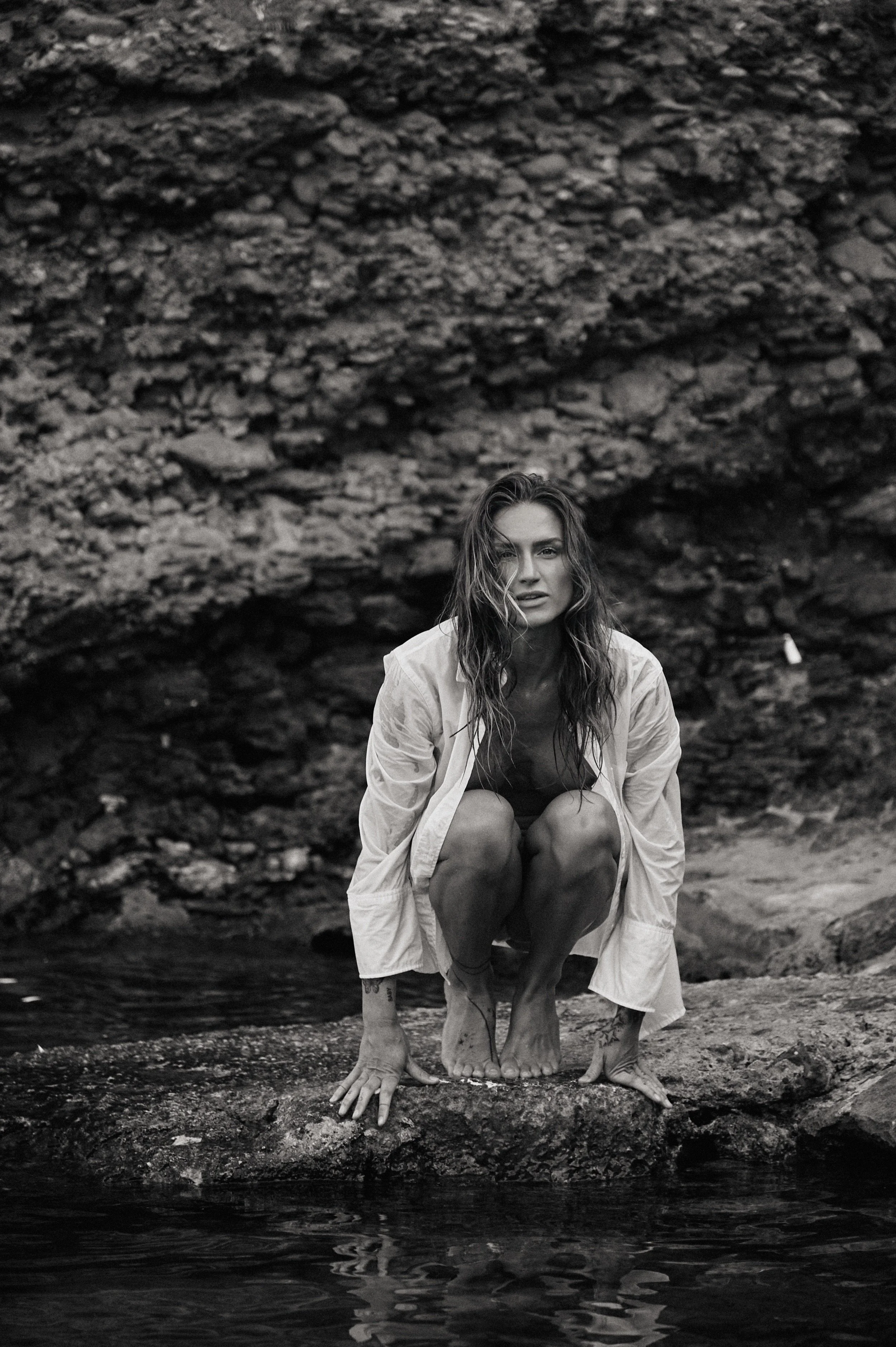 A woman crouching on a rock near water, with a rocky background, in black and white.
