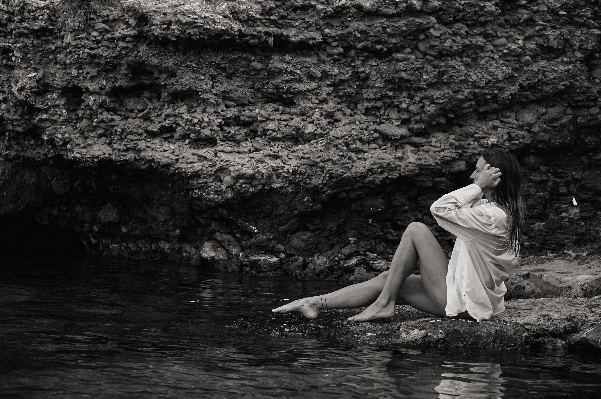A woman sitting on a rocky shore, partially in the water, adjusting her hair, with a rugged cliff in the background in black and white.