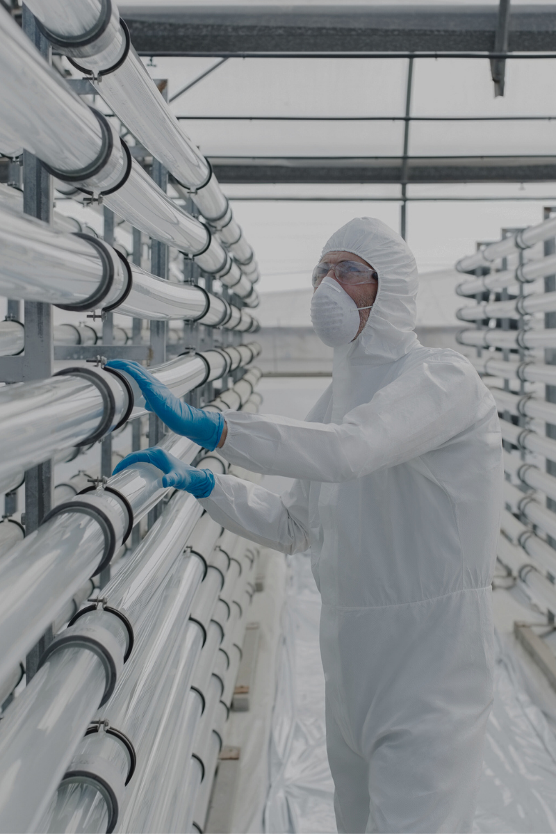 A scientist or technician wearing a protective suit, goggles, and a face mask working with large transparent tubes in a laboratory or industrial facility.