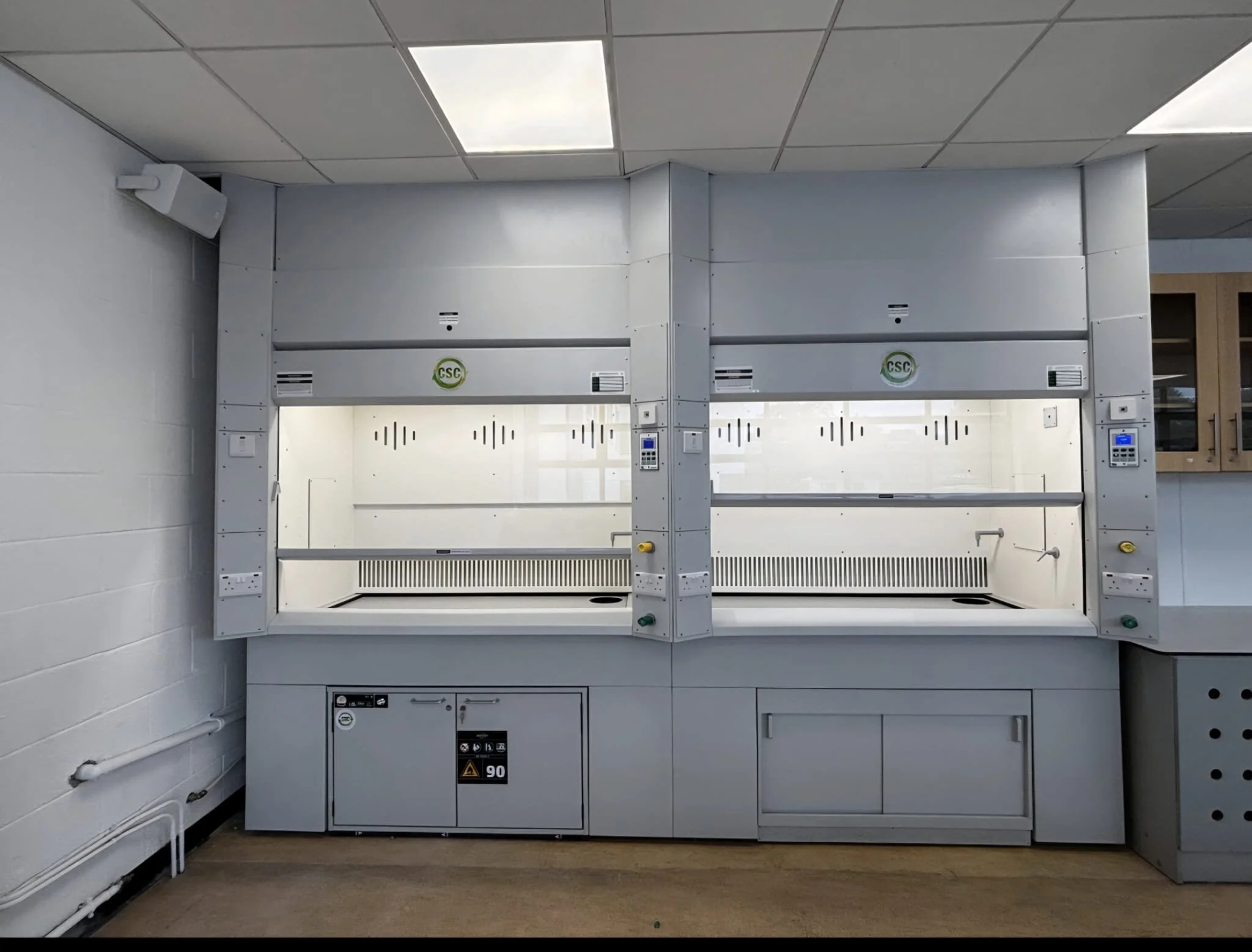 A clean, empty laboratory safety cabinet with glass front doors and control panels on the sides, situated against a white wall in a laboratory setting.