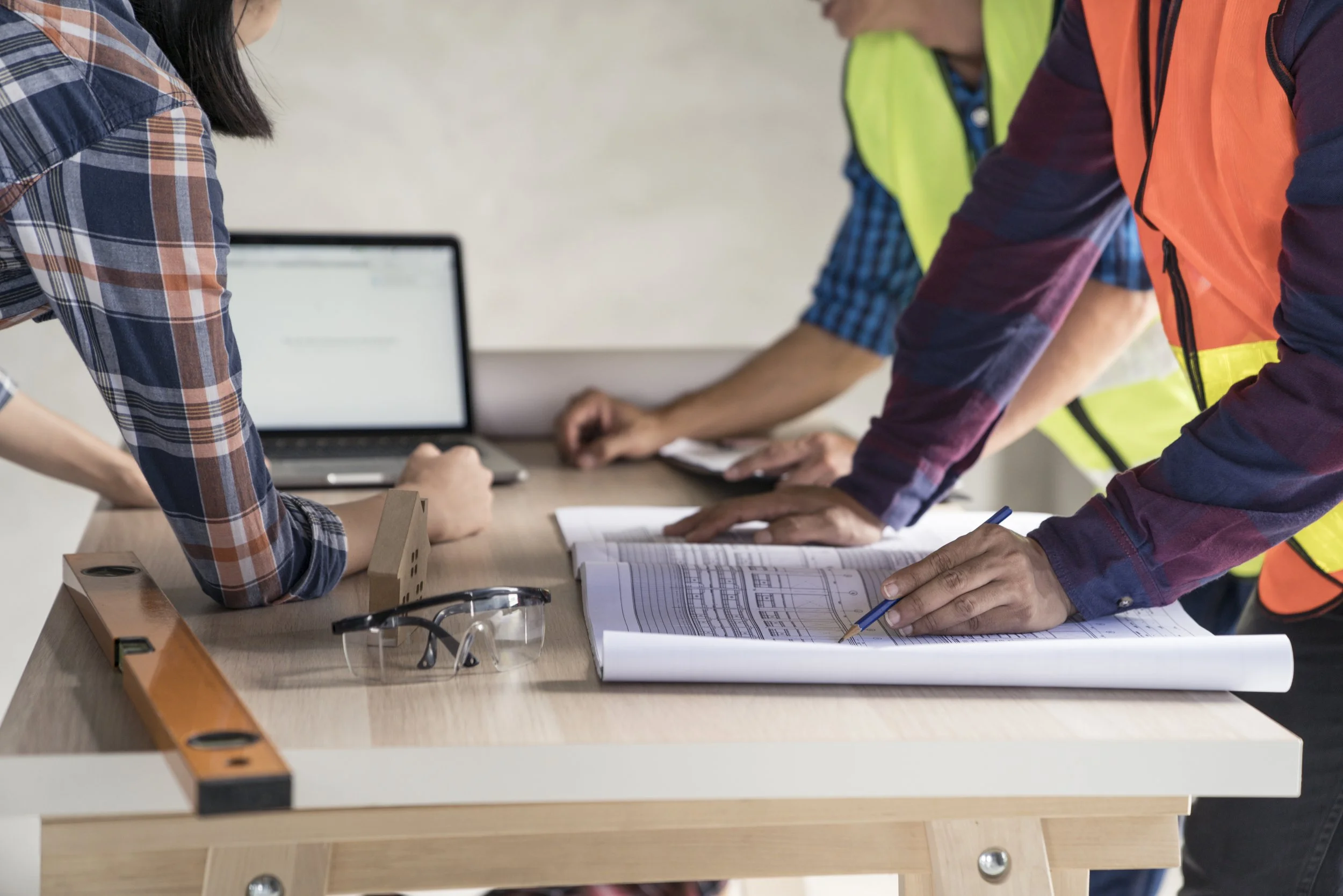 Three construction workers reviewing blueprints on a table with a laptop, level, and safety glasses.