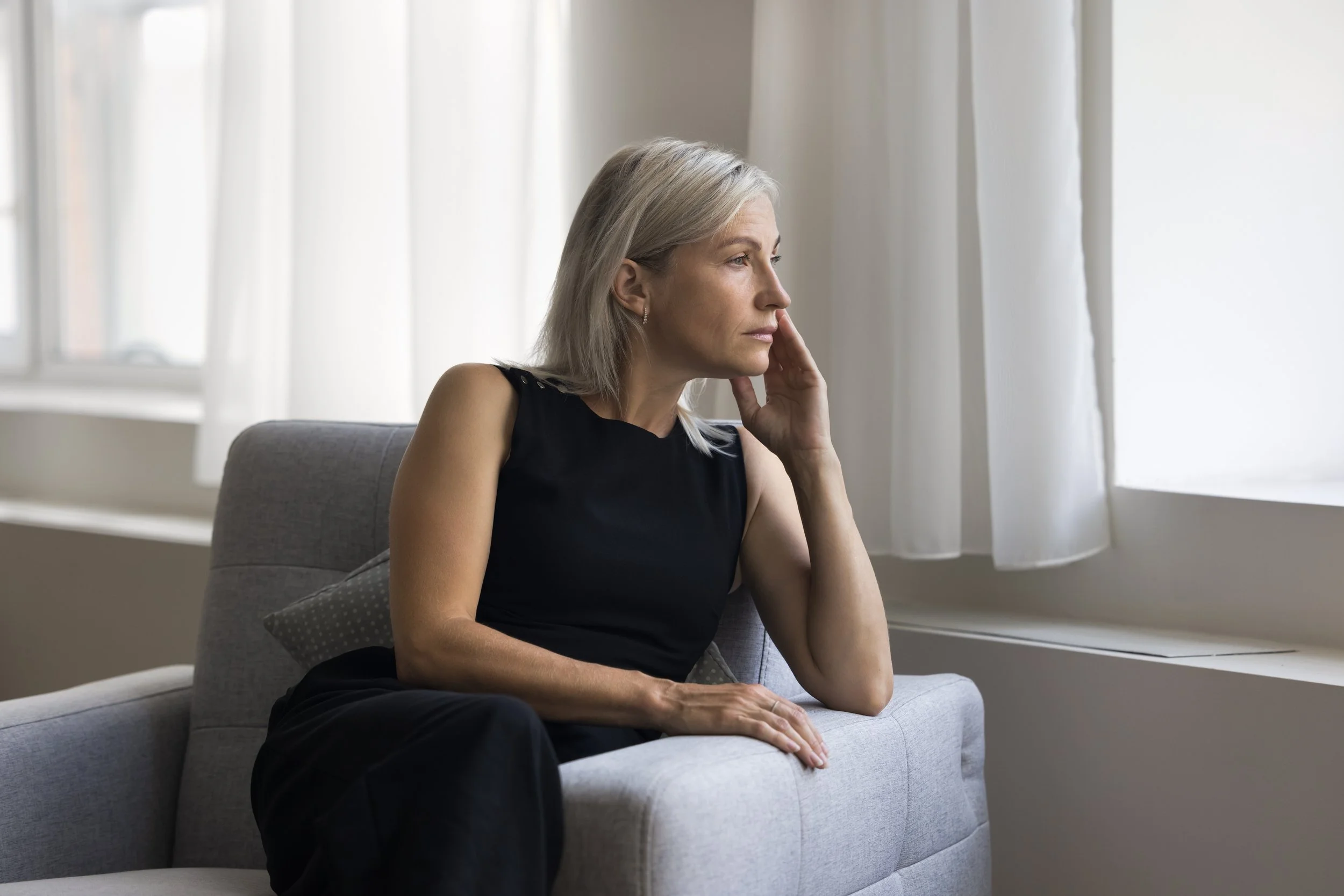 A woman with gray hair sitting on a gray sofa, gazing out a window with white curtains, in a contemplative pose.